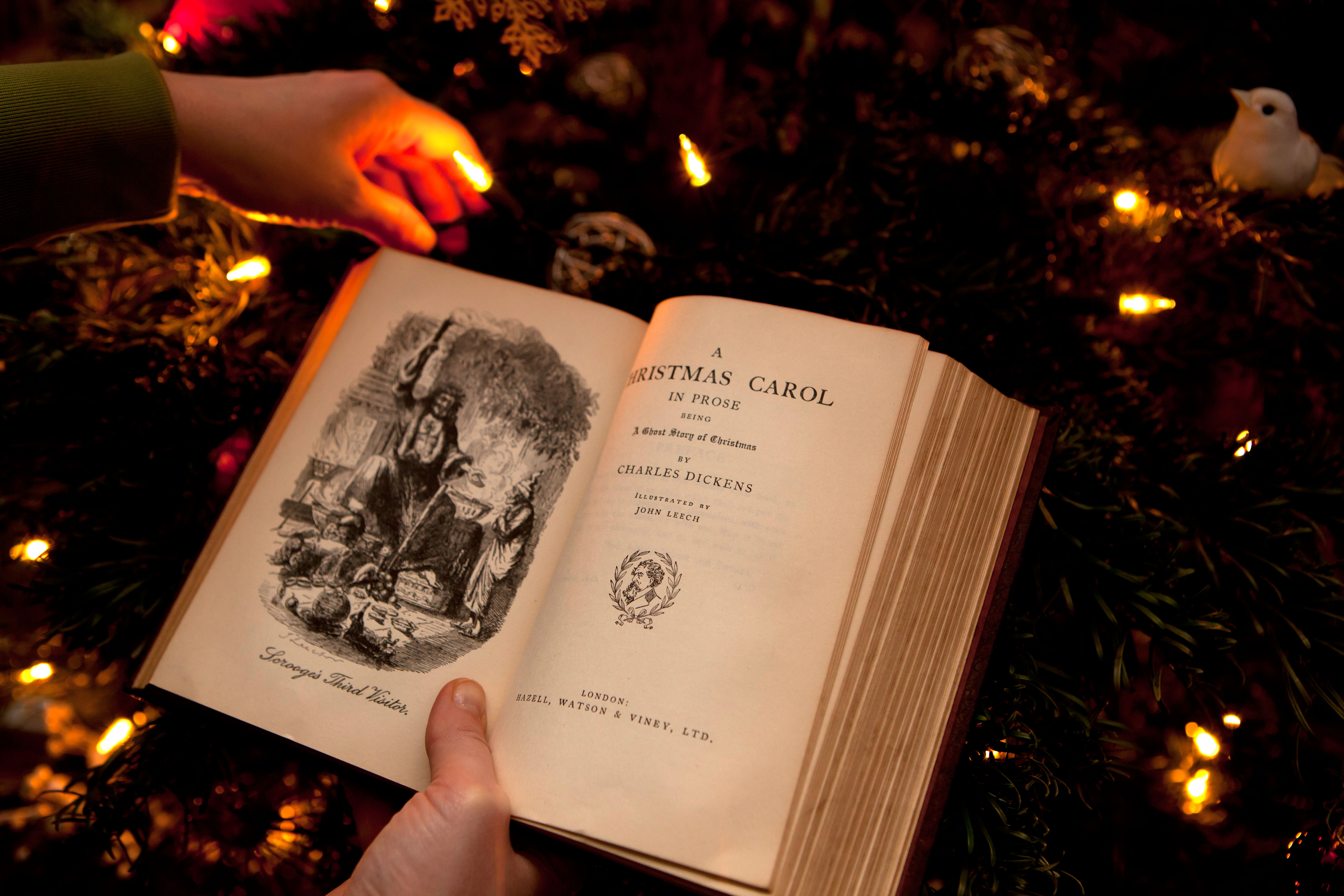 A hand holding a copy of A Christmas Carol in front of Christmas tree lights