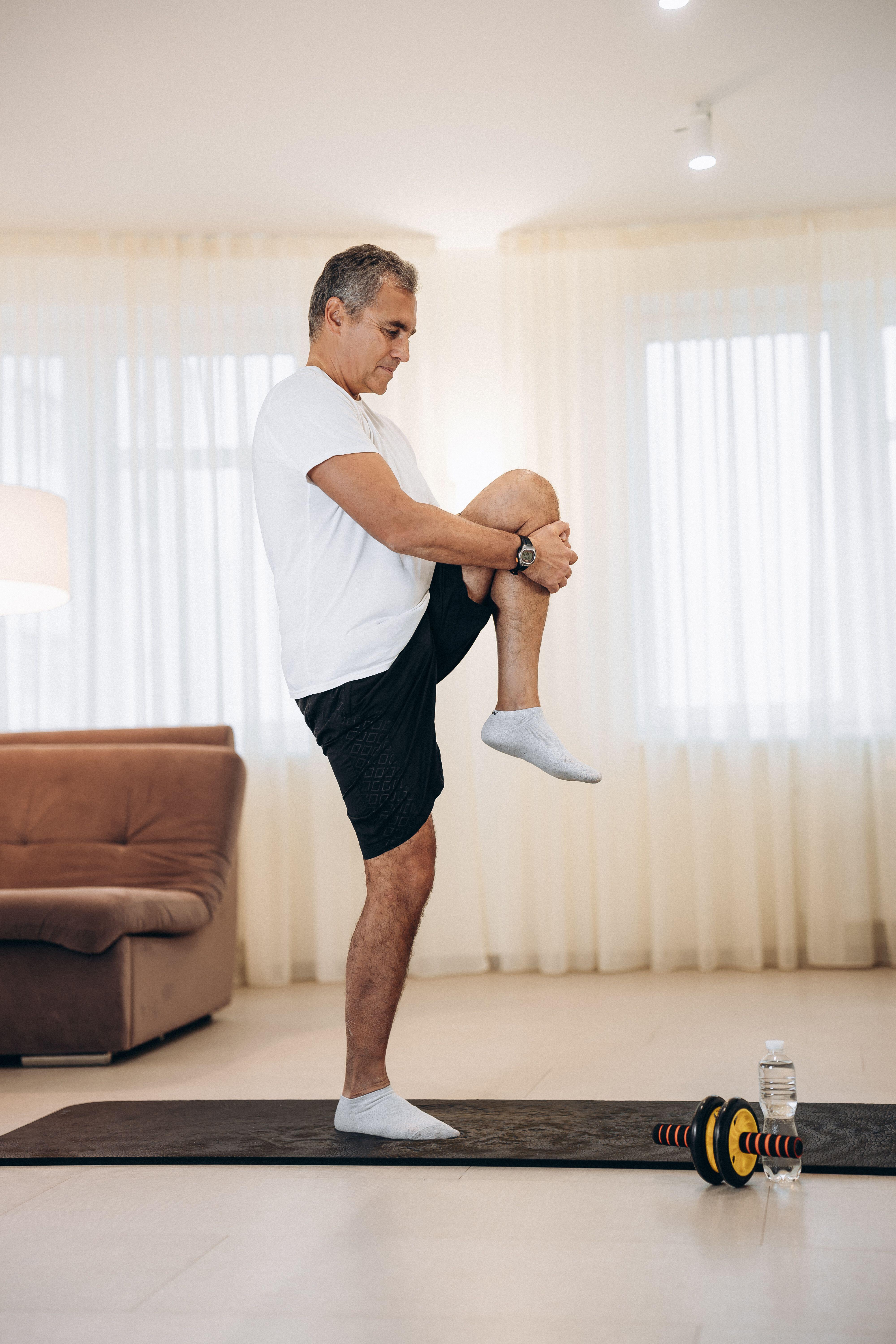 Mature man standing on one leg and stretching another in a living room next to a yoga mat and some weights