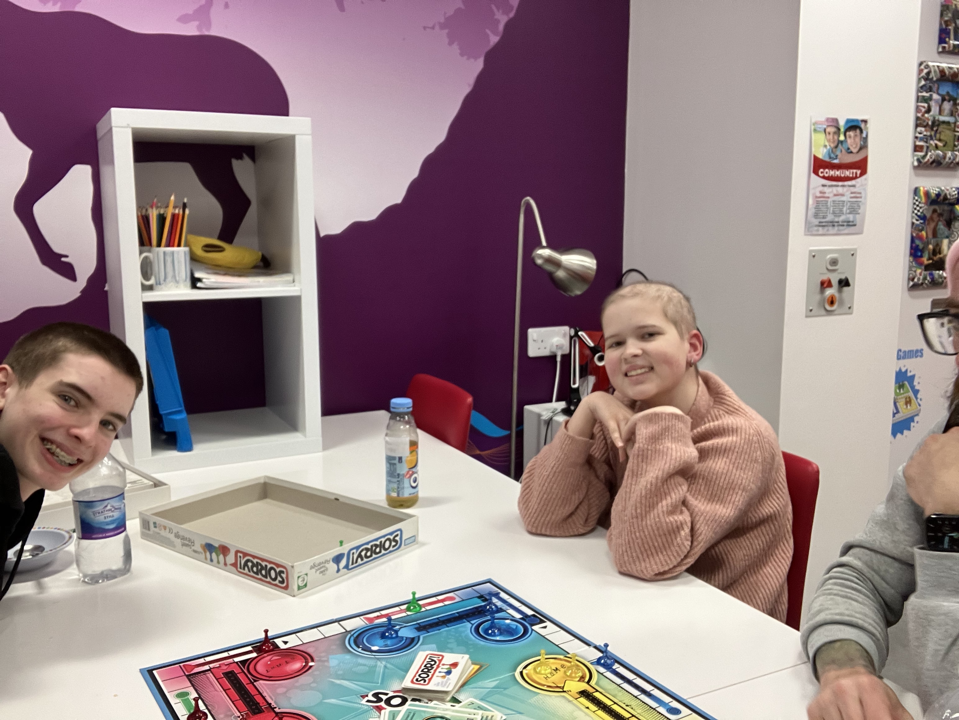 A group of three young people sitting around a table with a board game in the centre. The background features a purple wall