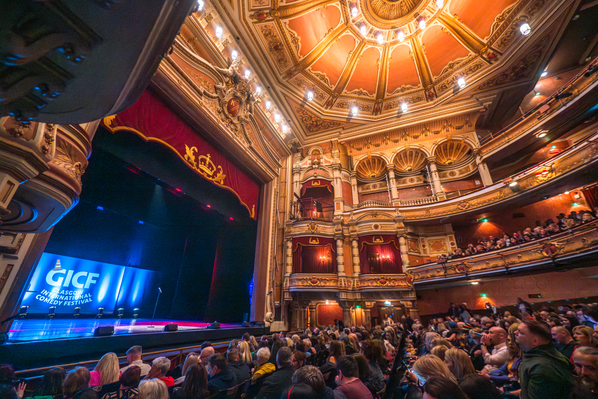 A shot of the lavish interior of the King's Theatre, looking up towards the richly decorated boxes and ceiling from within the stalls, with the stage in view on the left.