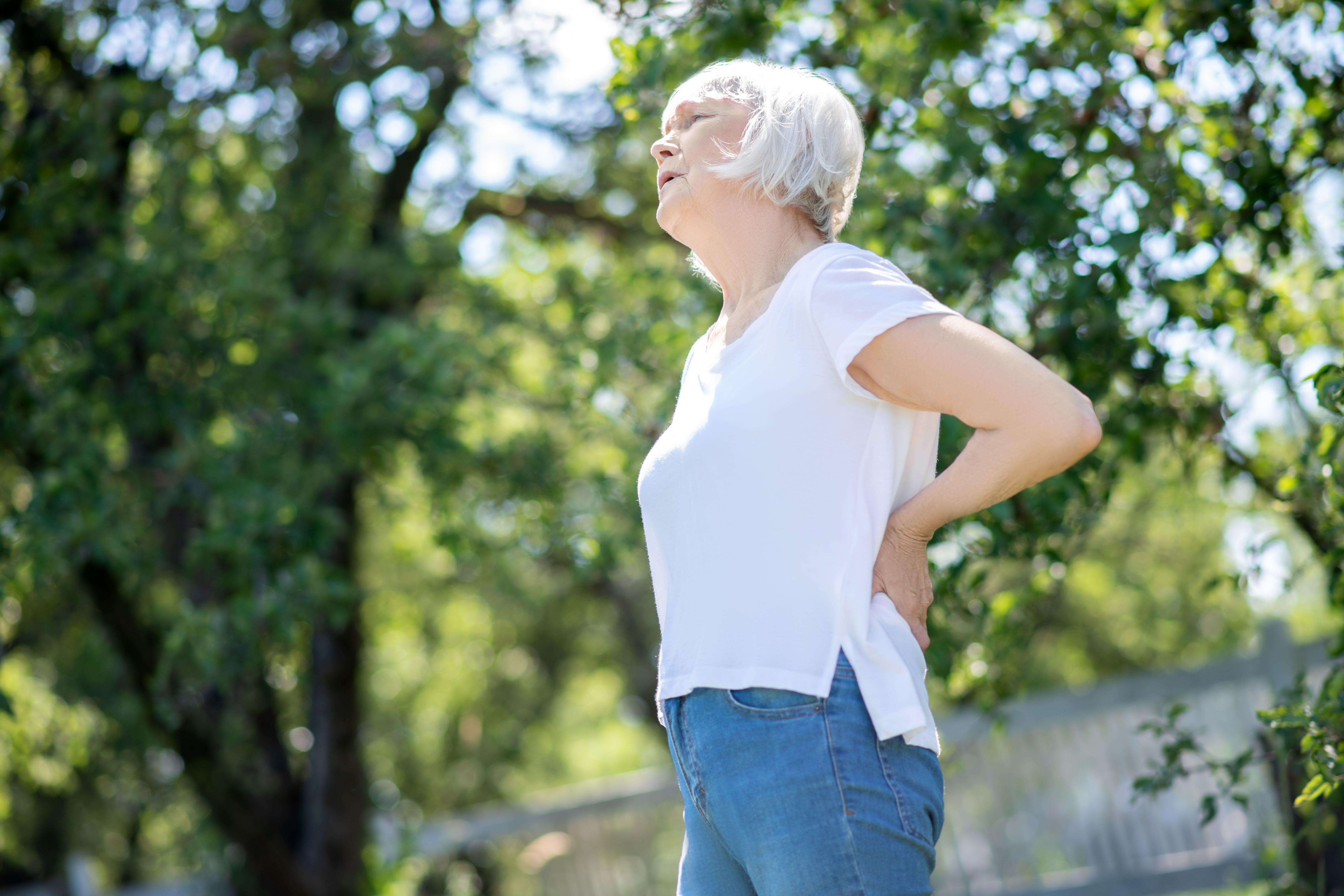 Senior woman wearing a white Tshirt and blue jeans standing outside suffering with back pain