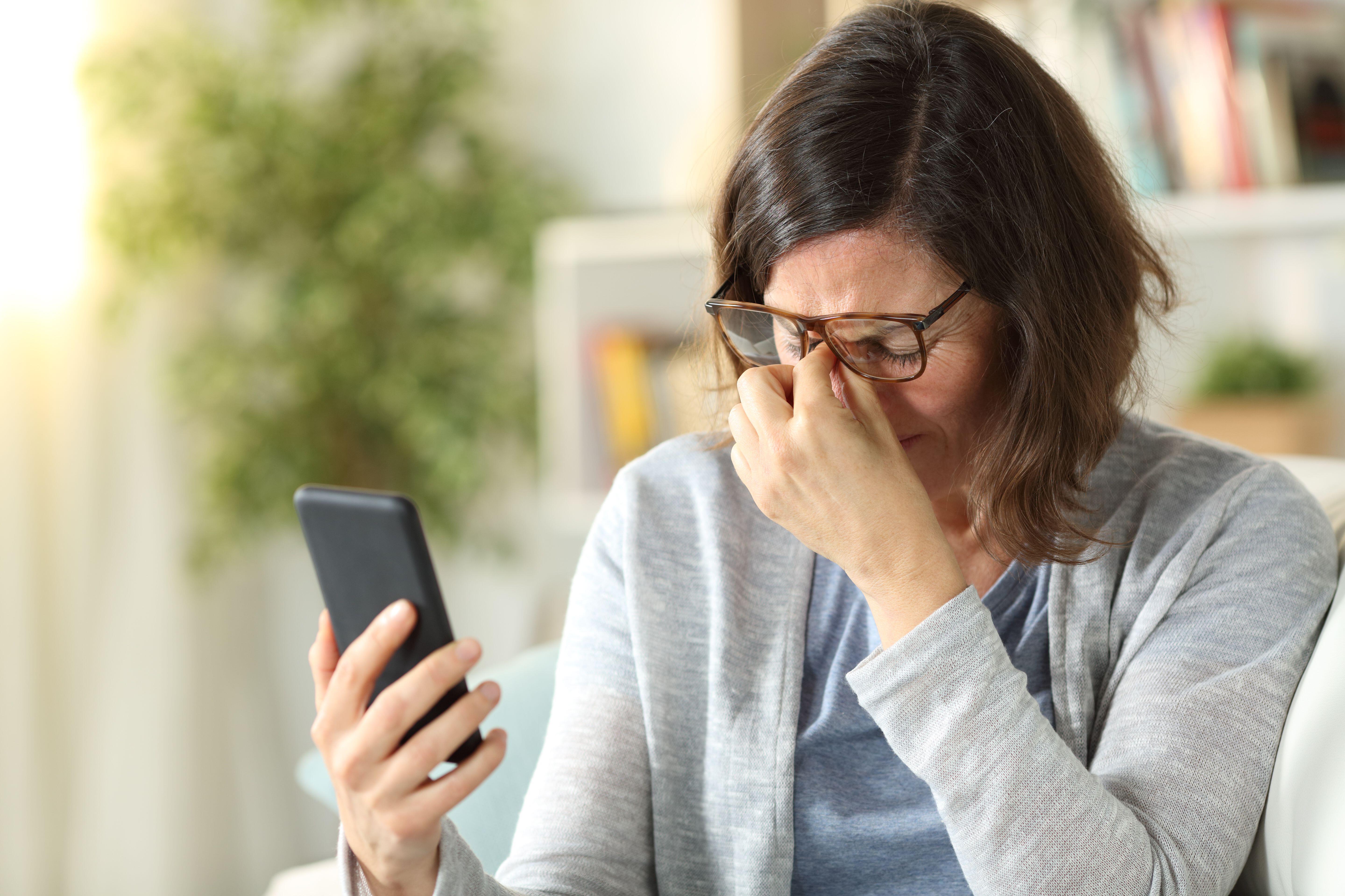 Adult woman, who is wearing glasses, rubbing her eyes and experiencing eyestrain after staring at her phone 