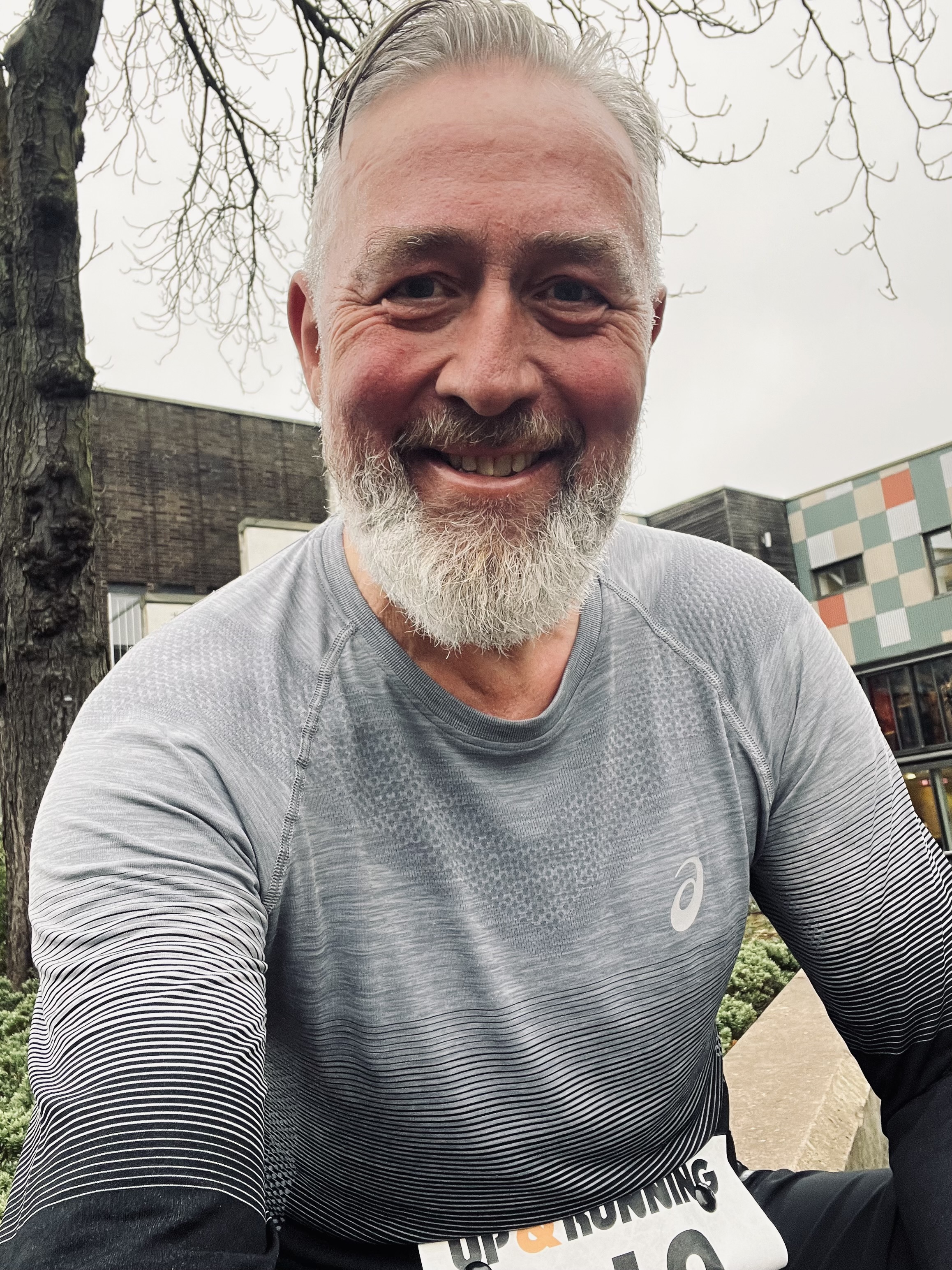 Ian Massey smiling at the camera while wearing a grey and black Asics long-sleeved running top and a race number