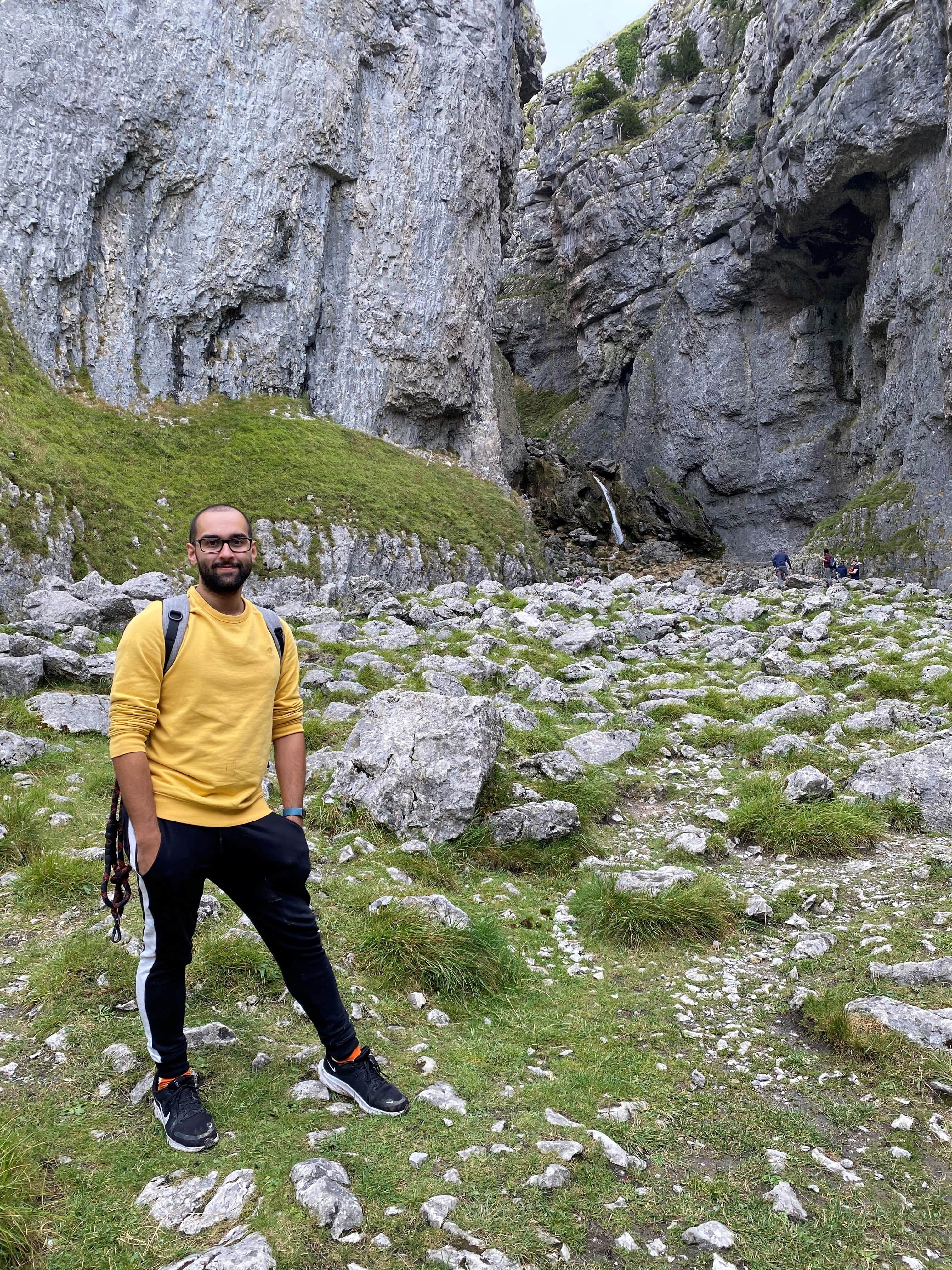 Michael Singh wearing a yellow jumper, black trousers and black trainers in a rocky landscape