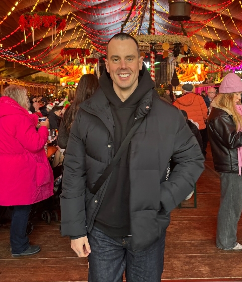 Sean Willers smiling while wearing a a black puffer jacker over a black hoodie and jeans 