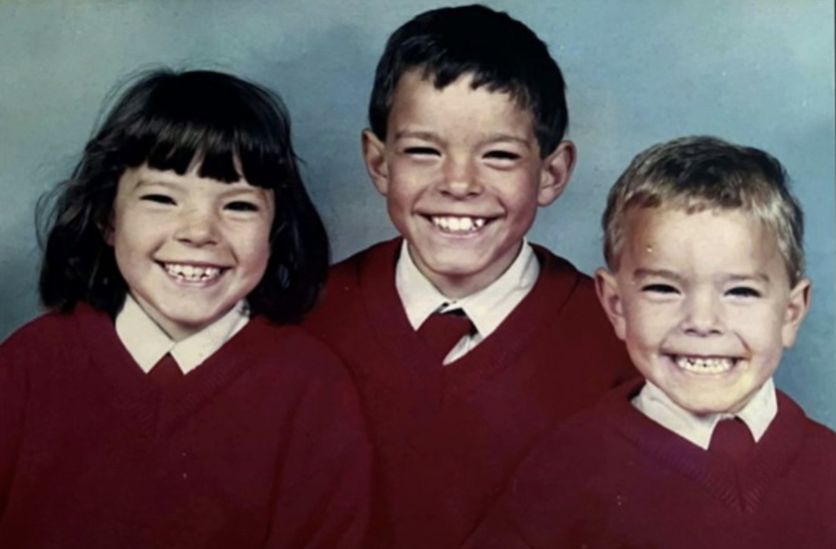 Three children in red school jumpers and white shirts, smiling at the camera