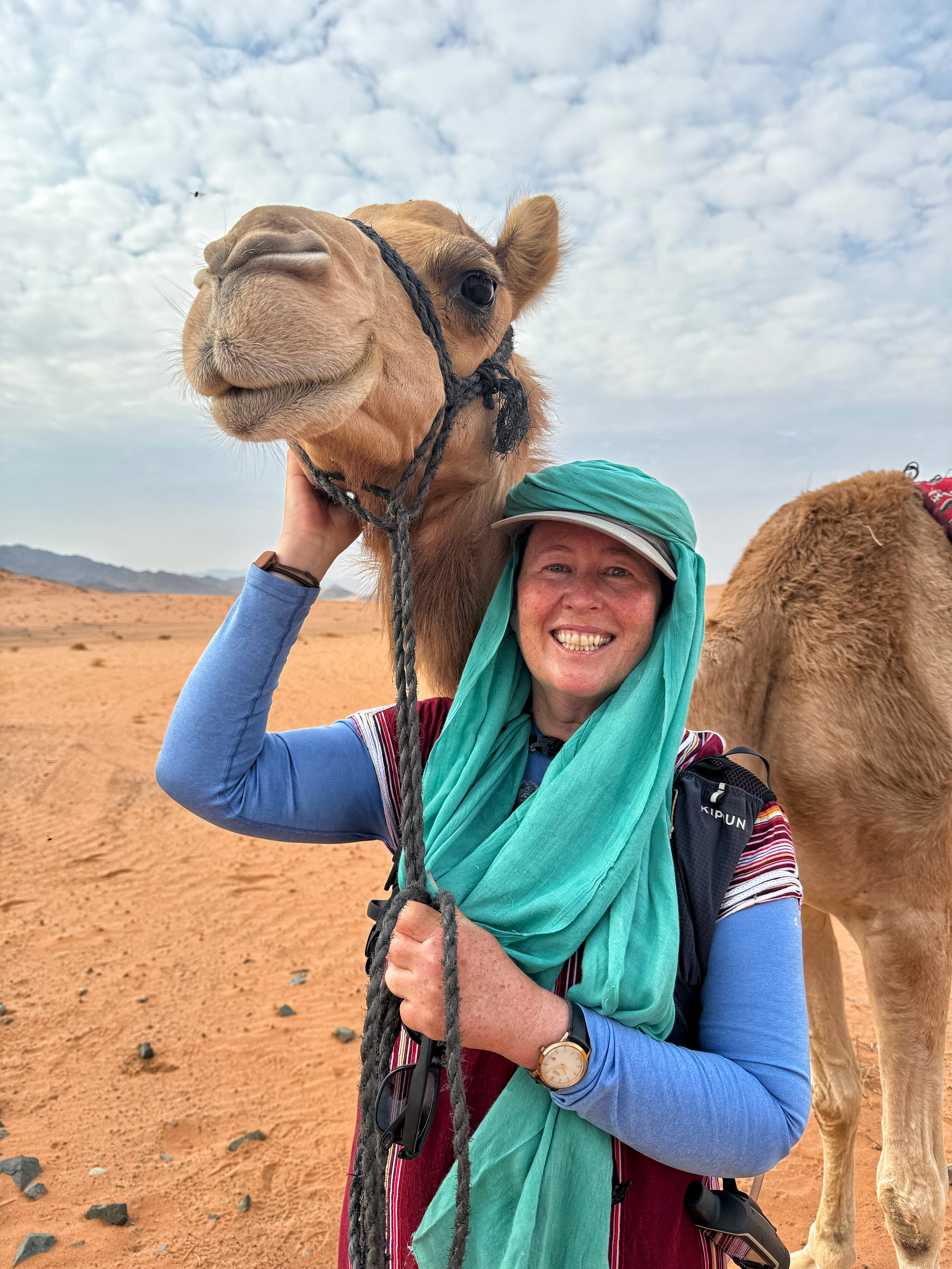 Alice Morrison smiling, wearing a green headscarf in front of a camel, one hand holding a rein and the other stroking the camel's face