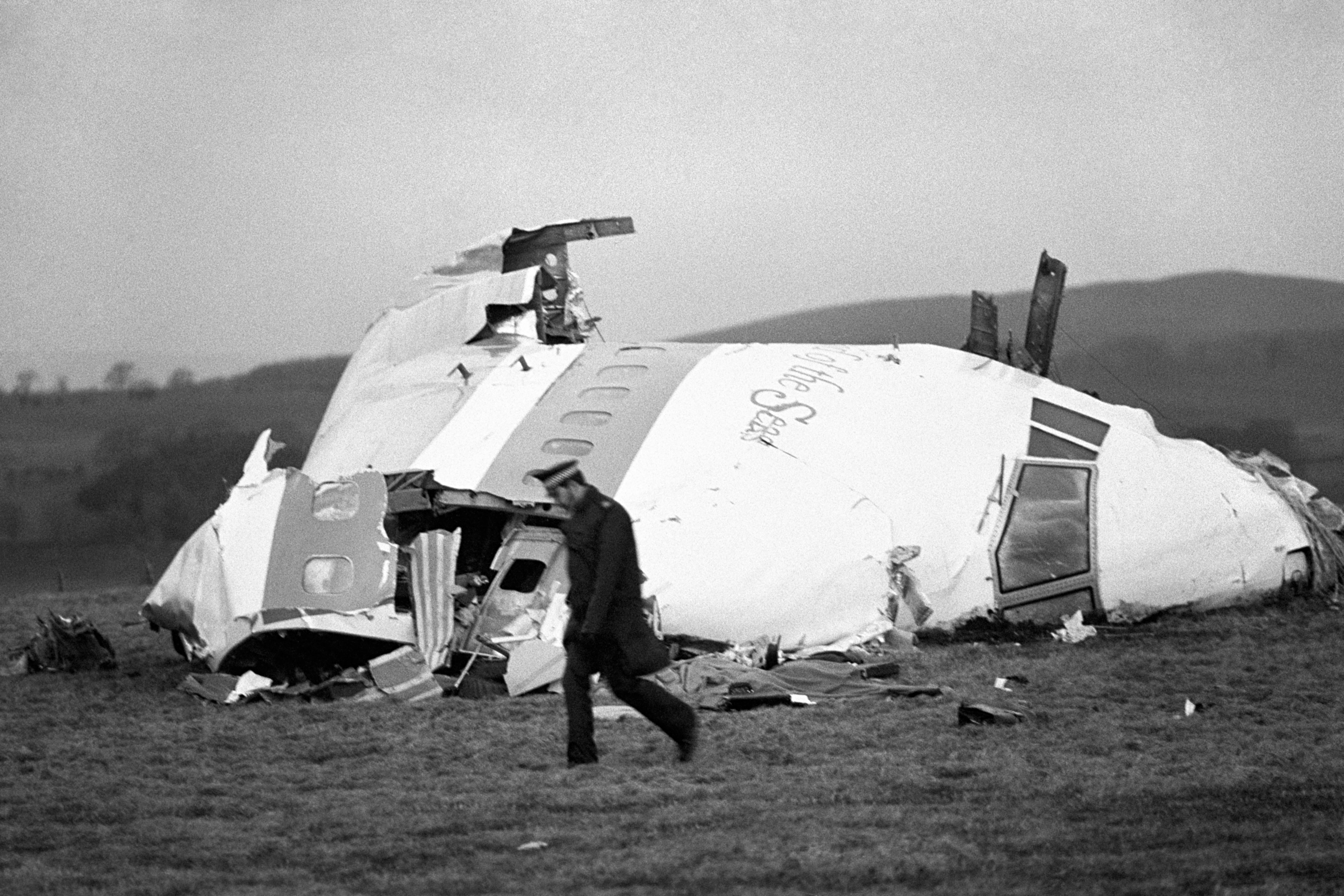 Black and white photo of a police officer walking in front of the nose cone of Pan Am flight 103 lying in a field