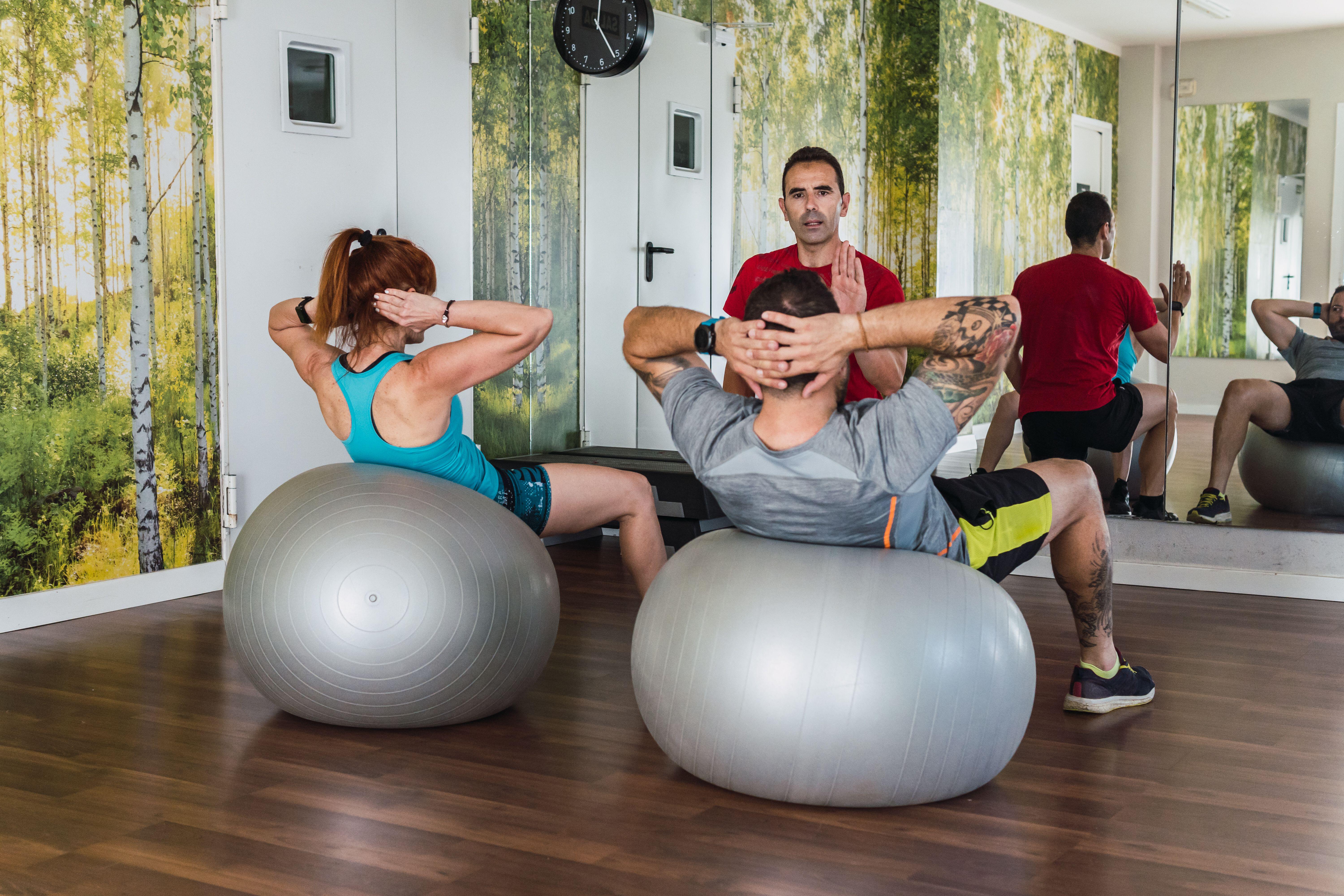 A personal trainer on bended knee coaching a man and a woman doing sit-ups on exercise balls in a gym