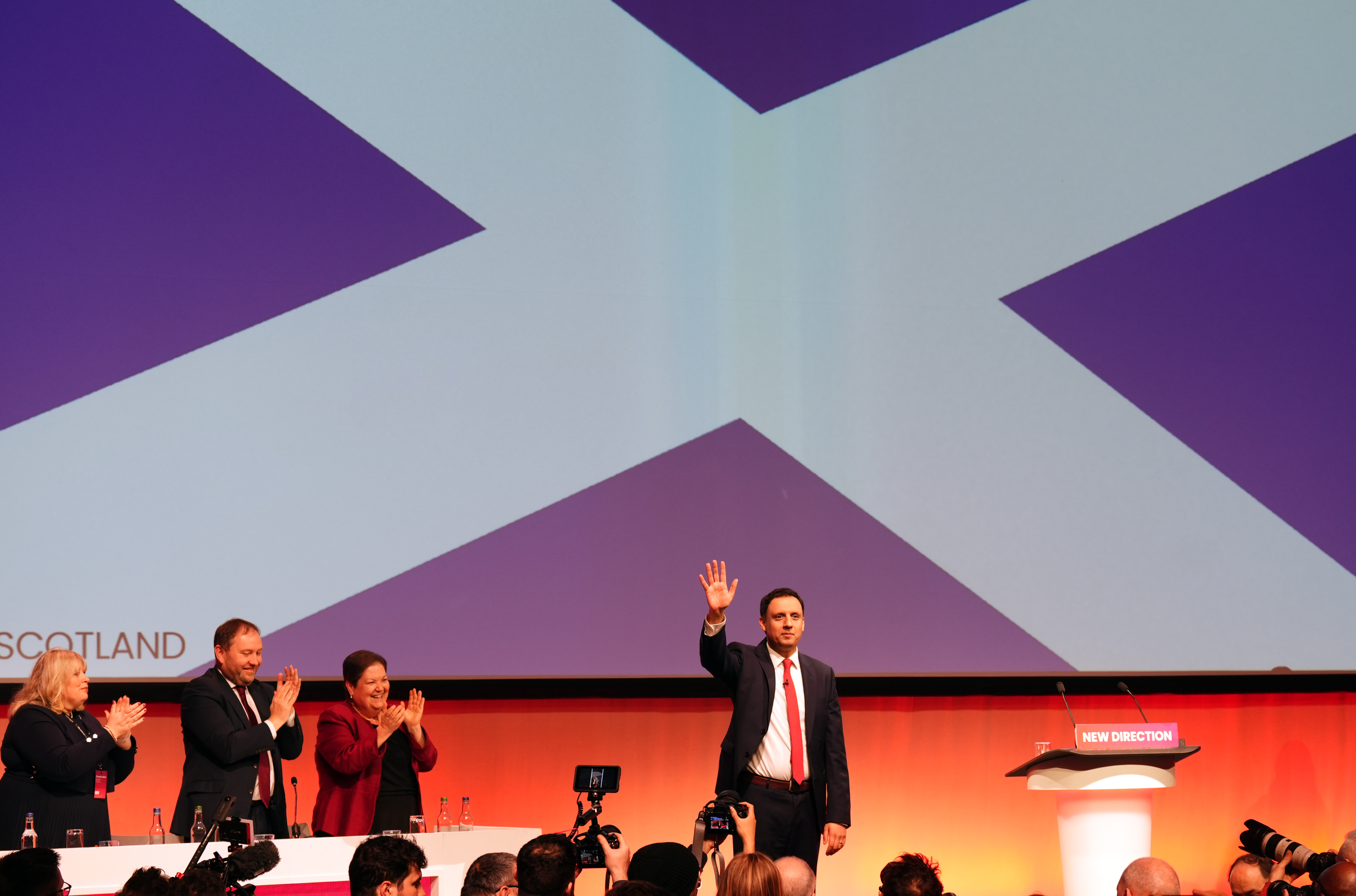 Anas Sarwar waves in front of a large Scotland flag, with senior Labour officials clapping from one side