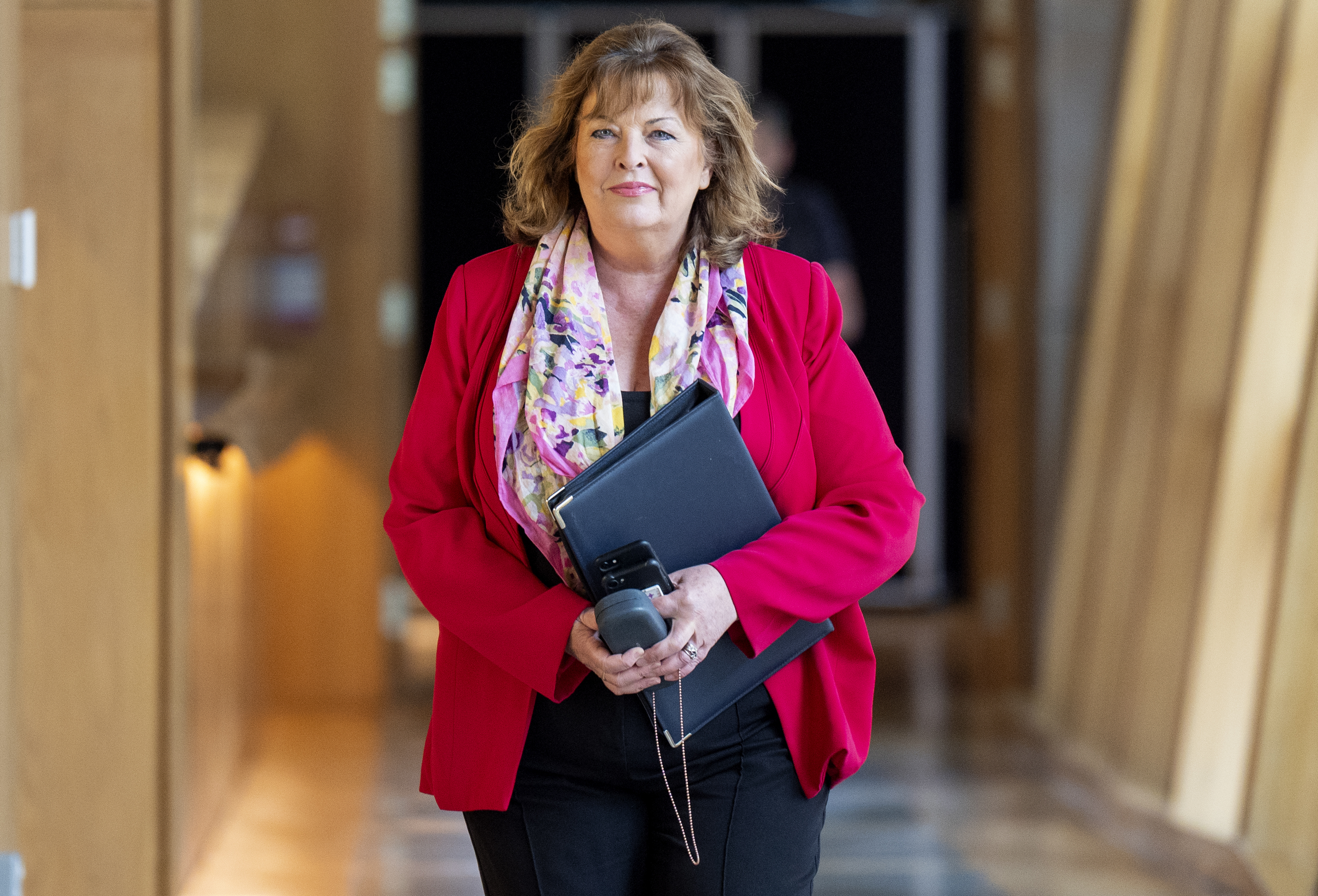 Fiona Hyslop smiling, wearing pink jacket and floral scarf, walking through Parliament