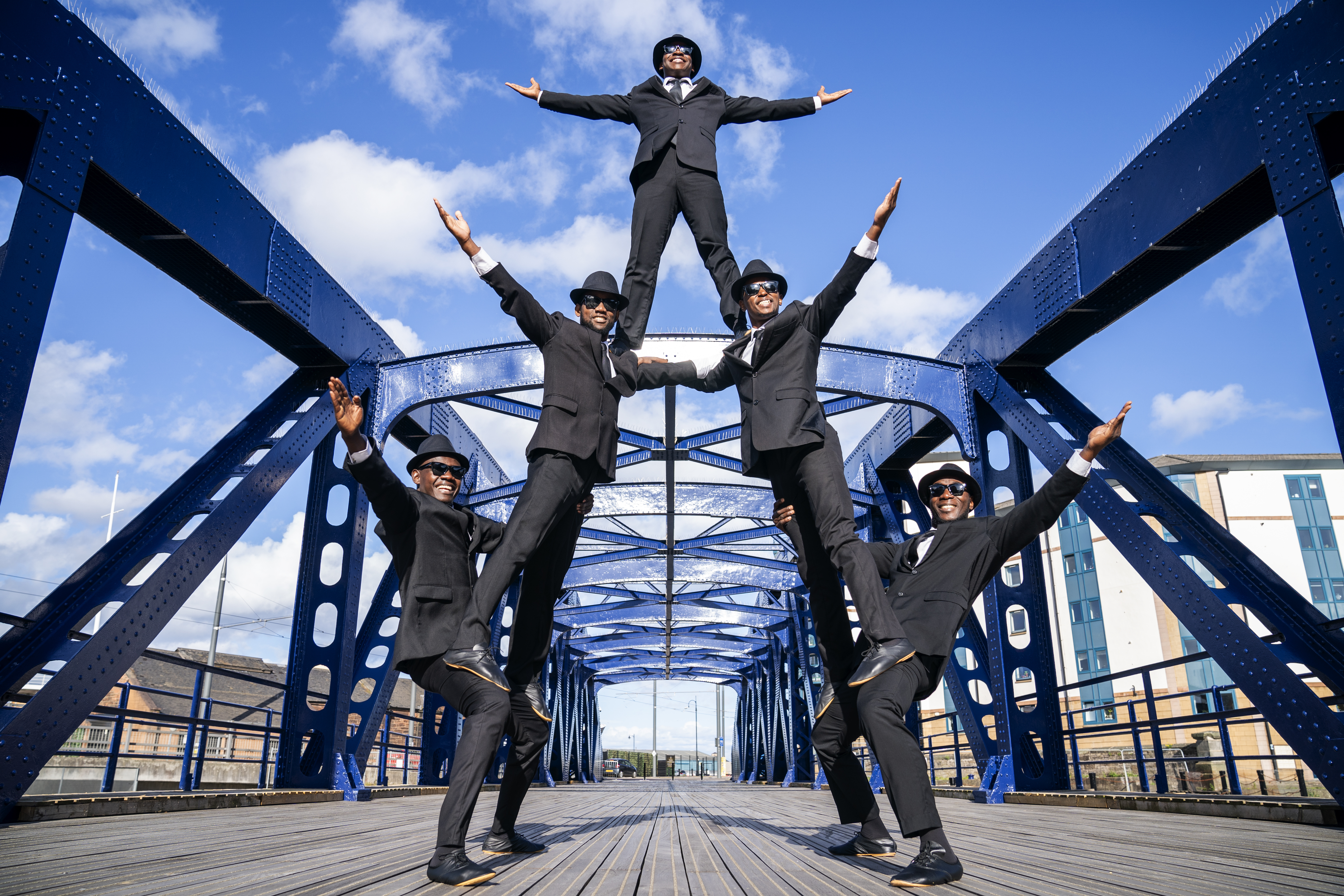 Five performers dressed in suits and hats, balanced on each other on a bridge