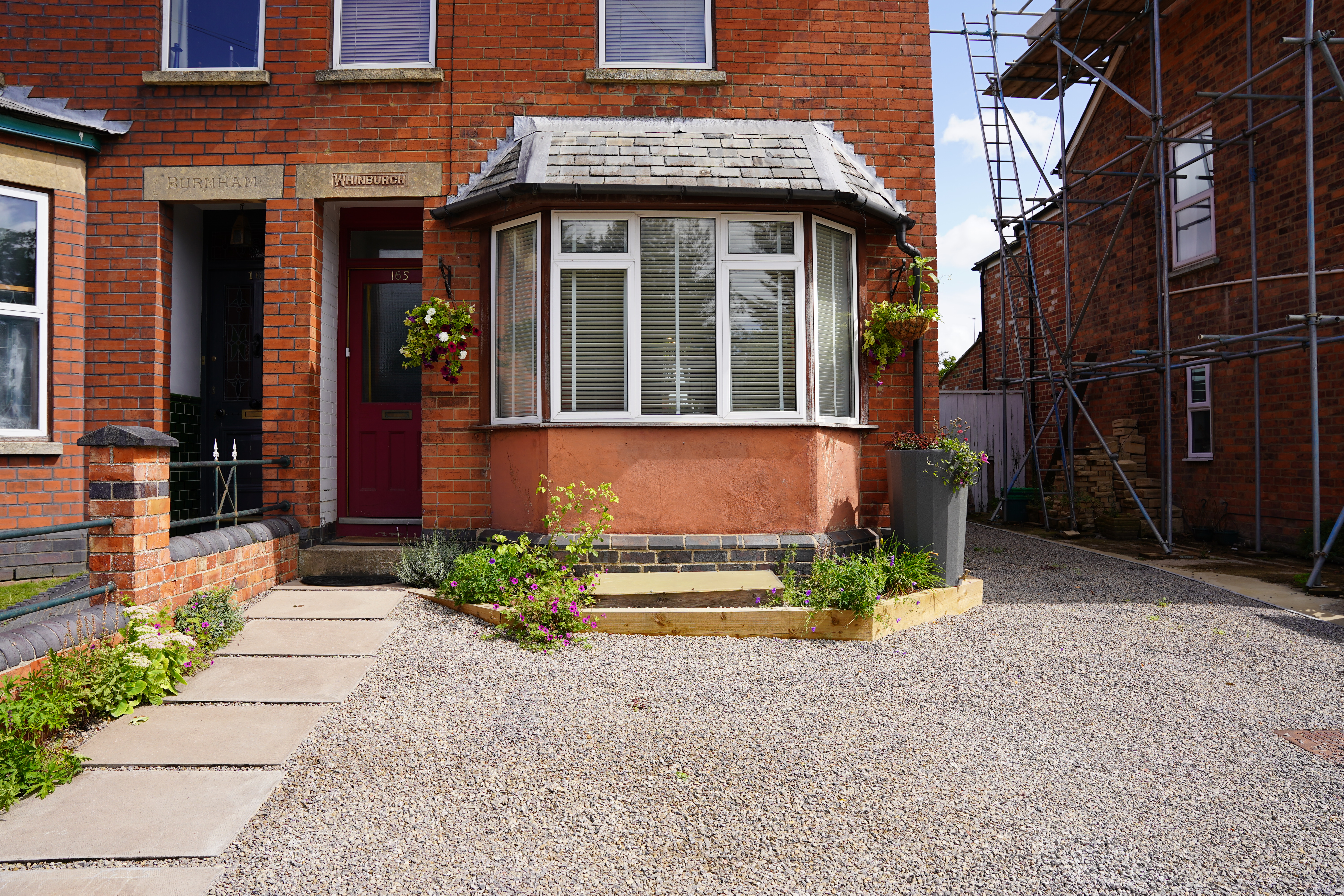 Gravel driveway with planting and water butt in front of semi-detached red brick house with bay window