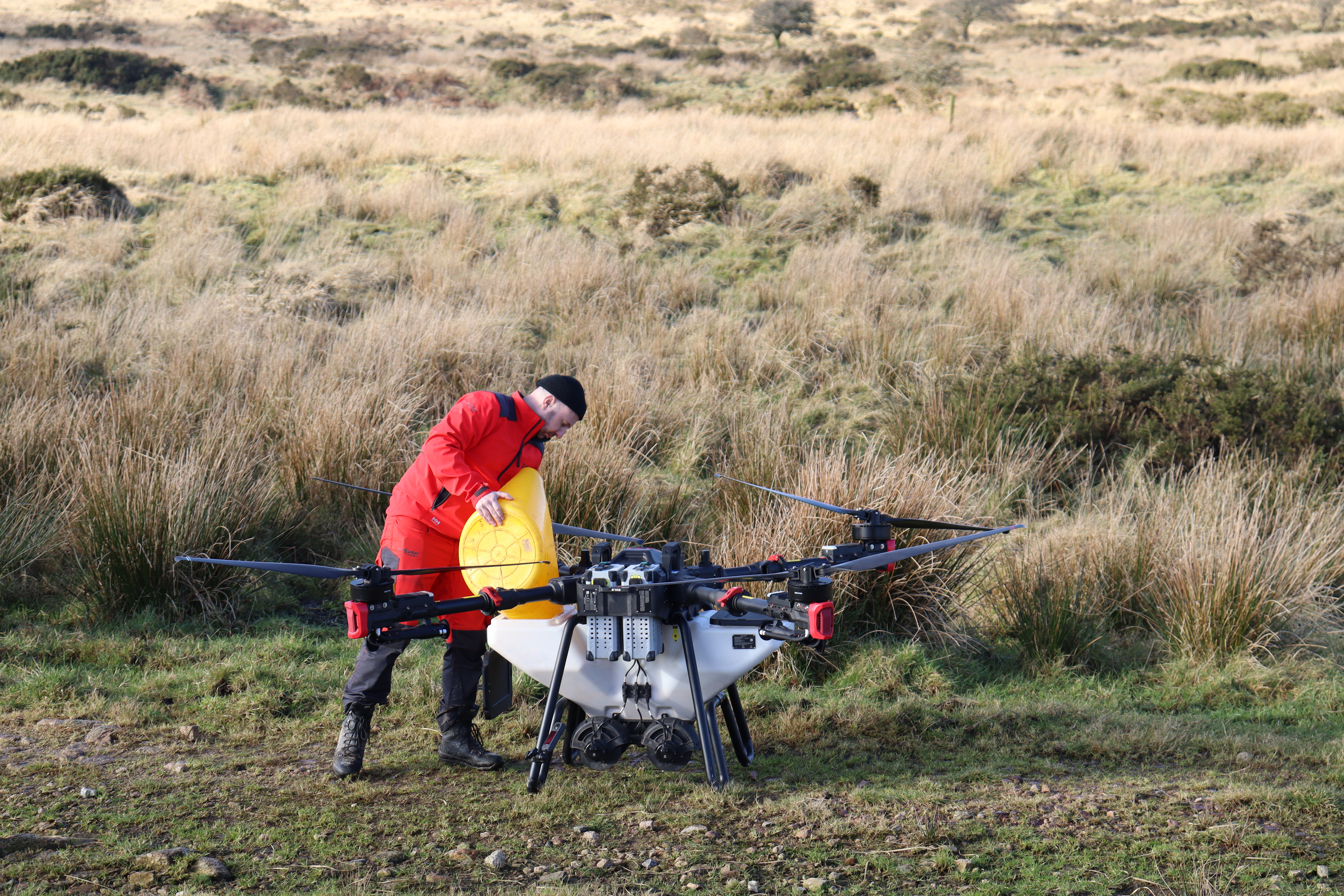 A large drone on the ground is being filled by a man in red waterproofs using a large yellow bucket