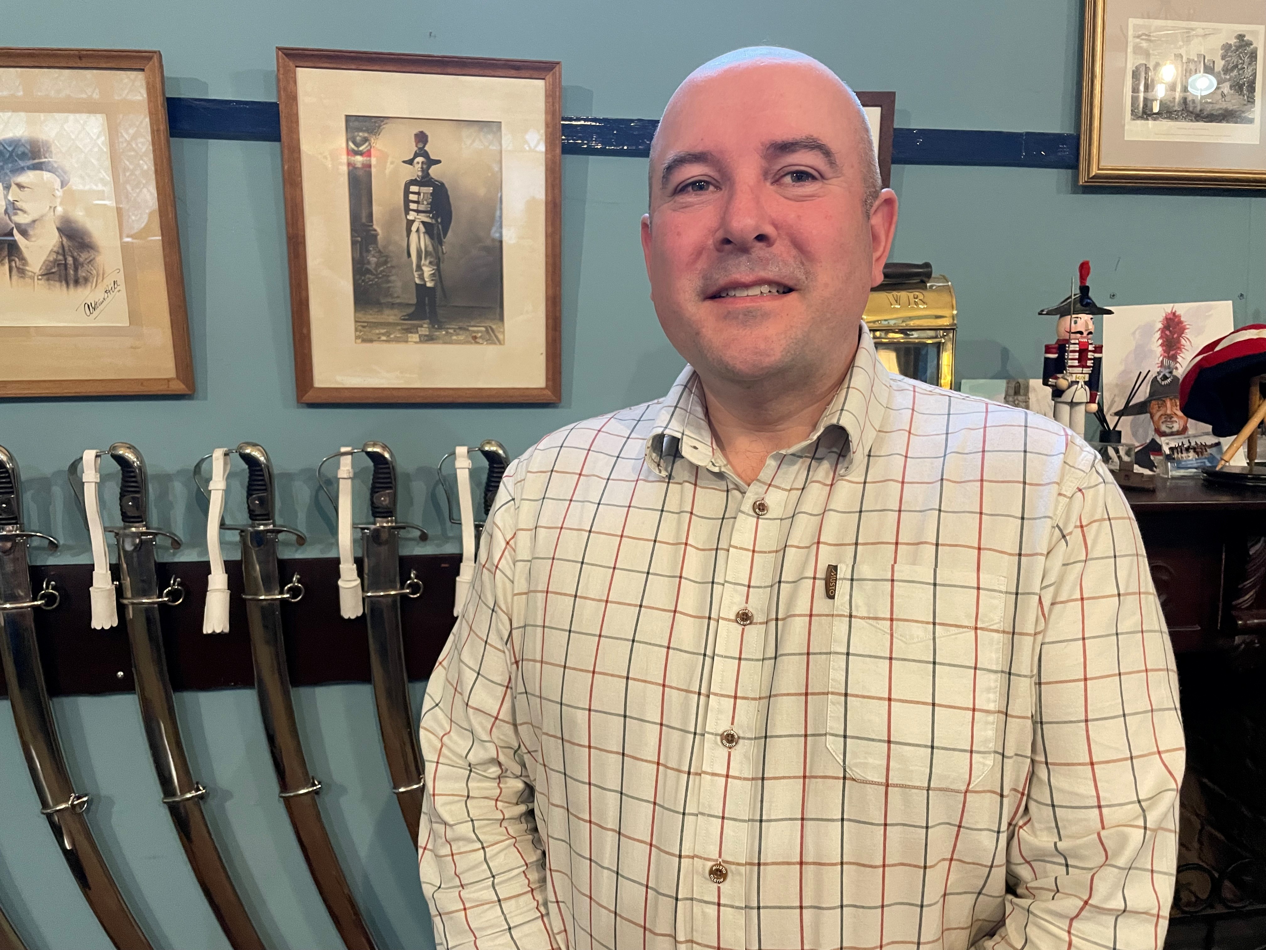 Andrew Carlisle smiling, with several ceremonial swords and historic photos on the wall behind him