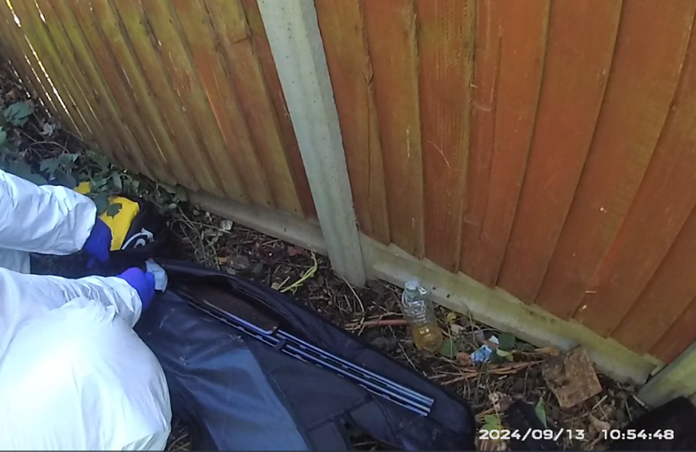 A forensics officer with a shotgun on ground next to a fence