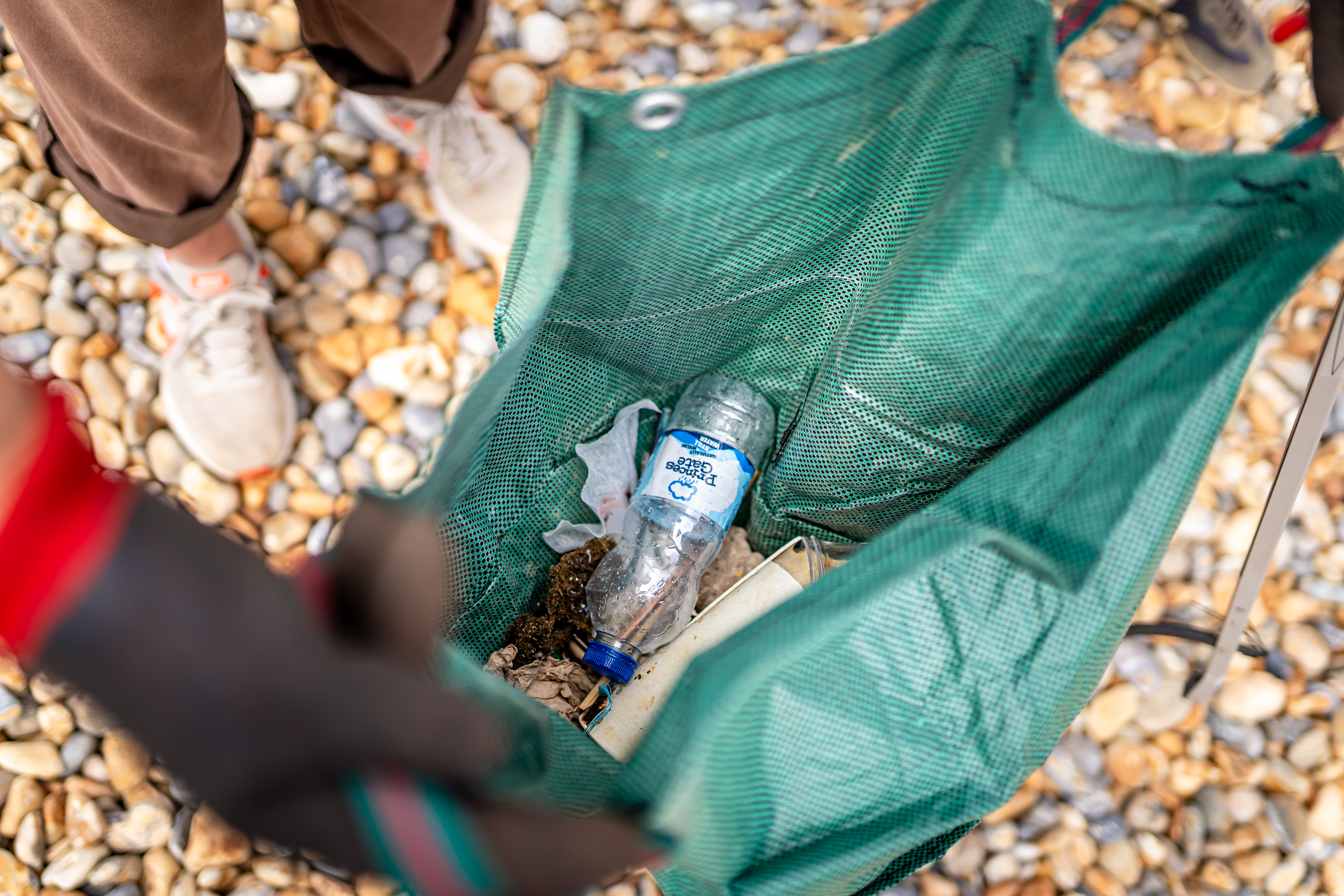 A green bag with plastic bottle and other rubbish is held open on a pebble beach