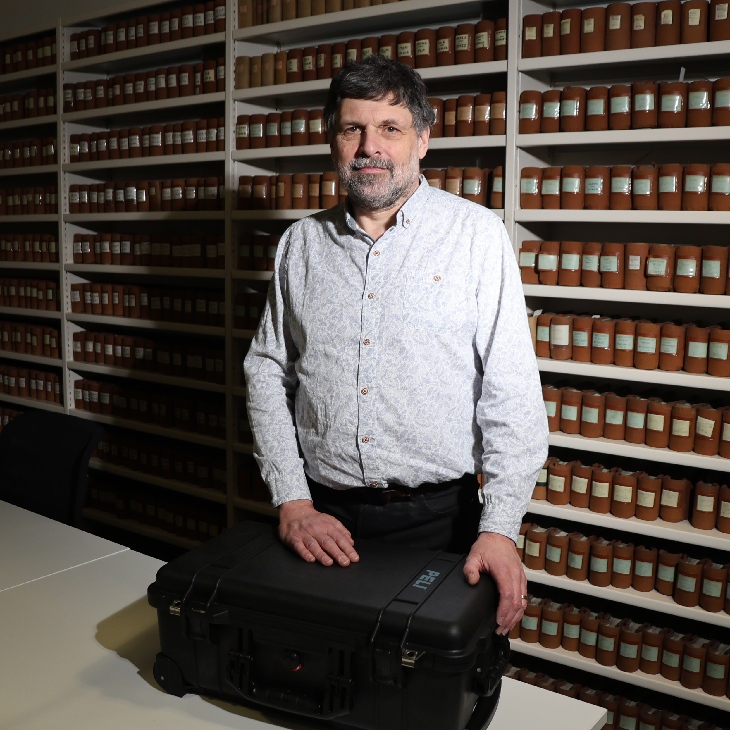 Neil Curtis of the University of Aberdeen standing in front of rows of shelves containing red objects, with his hands on a suitcase resting on a table