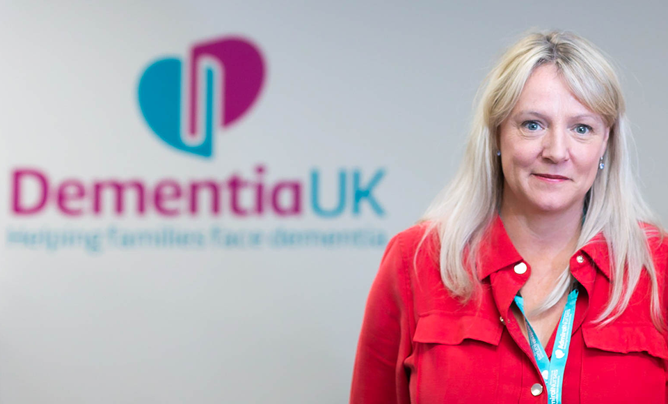 Helen wearing a red shirt and smiling standing in front of a sign for Dementia UK 