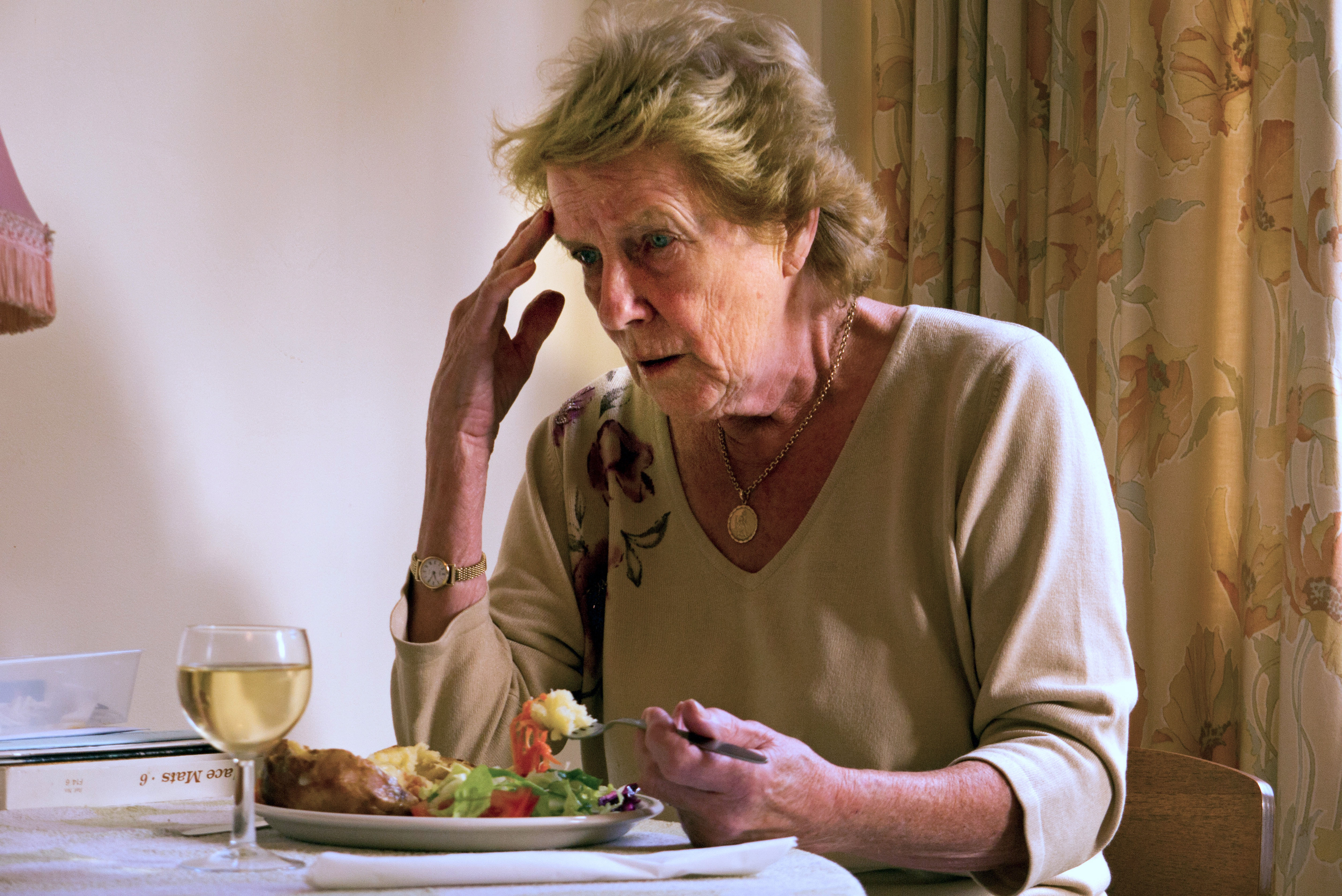 An elderly woman with Alzheimer's Disease sitting in her home eating a meal looking confused and worried.