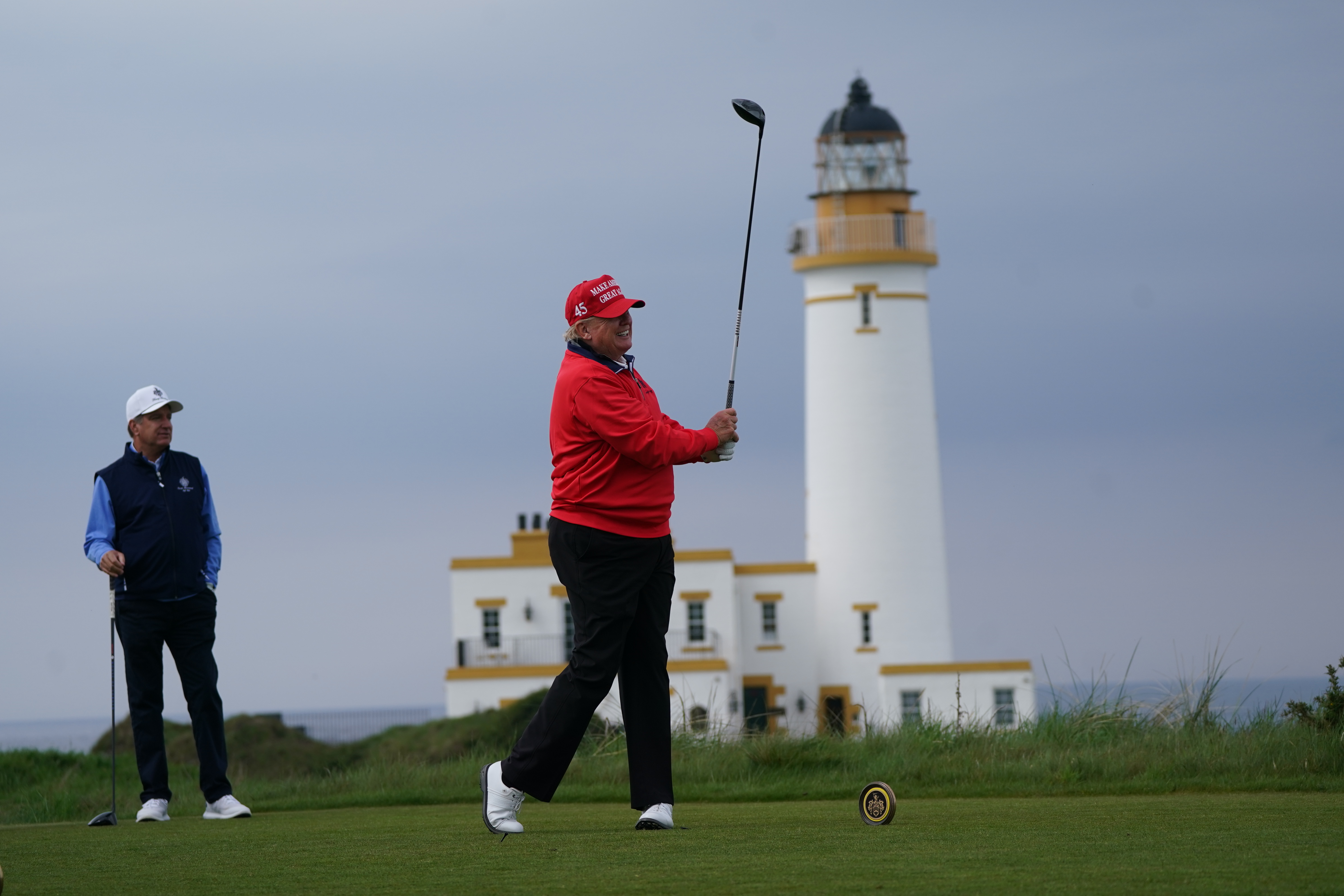 Donald Trump teeing off on a golf course, with a lighthouse in the background