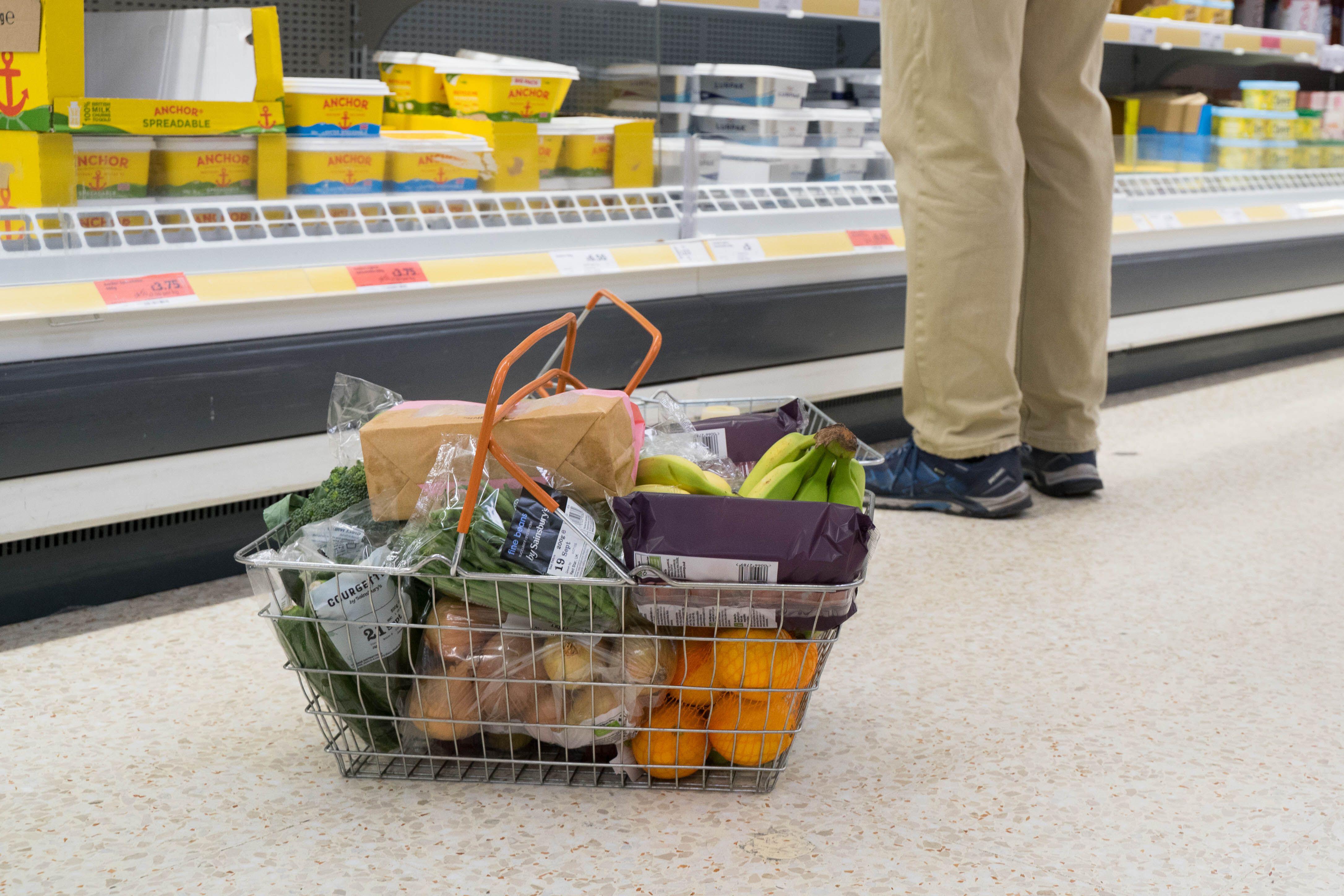 A shopping basket full of items on the floor of a supermarket, next to a shopper