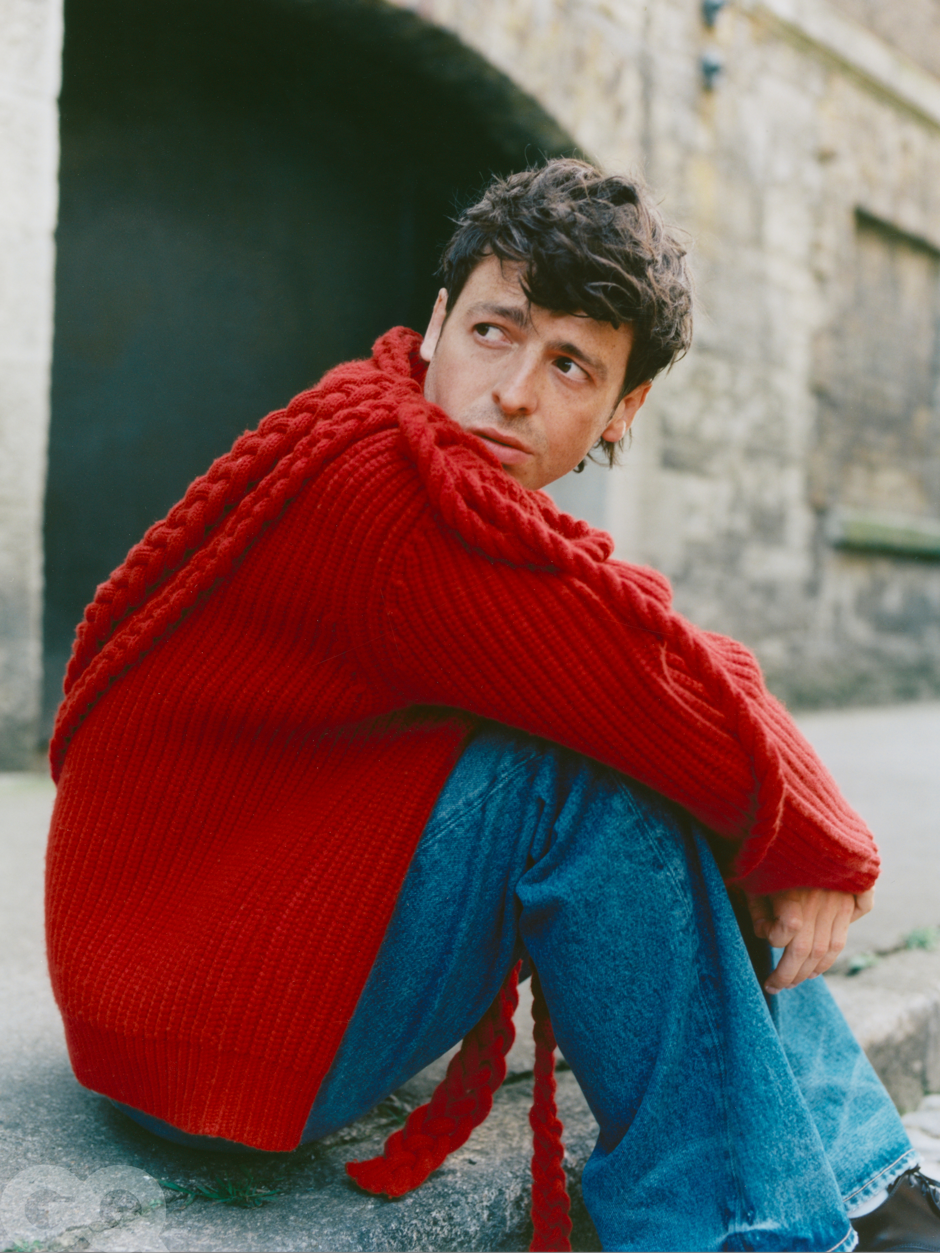 Anthony Boyle posing for a photoshoot, wearing jeans and a red jumper