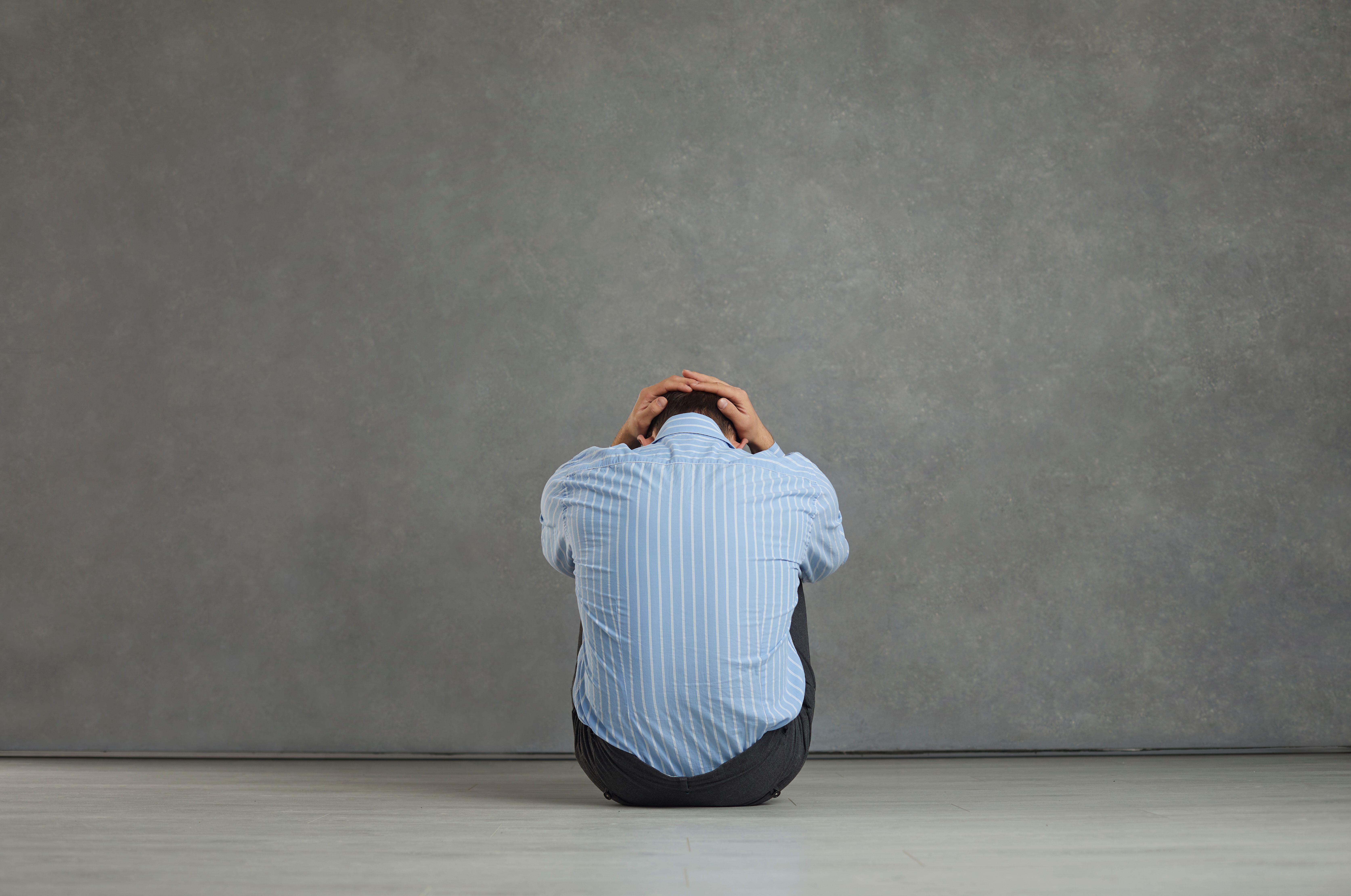 Back view of man sitting on floor, holding head, feeling distressed, frustrated and hopeless