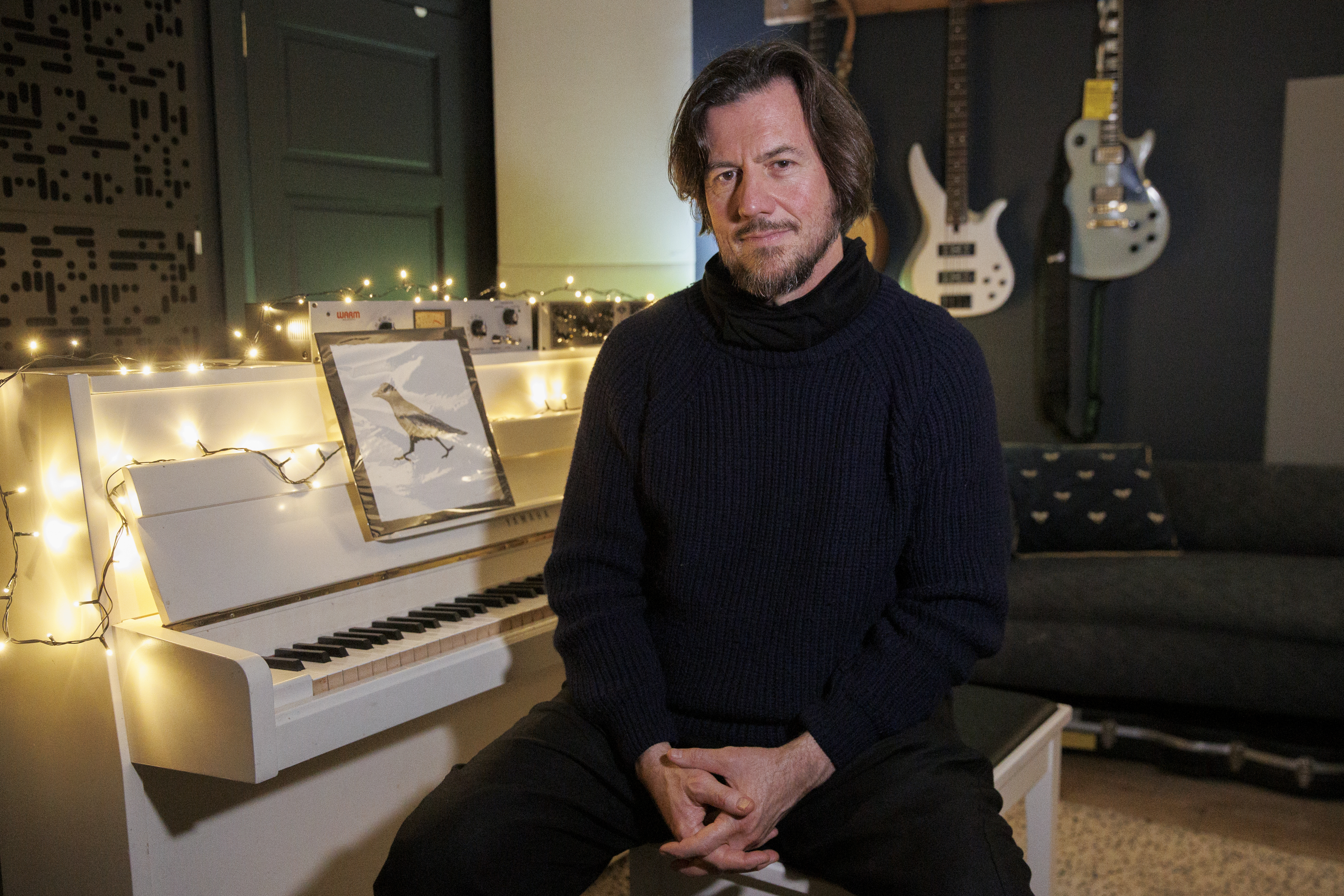 Robbie Boake pictured seated at a piano, with guitars hung up behind him