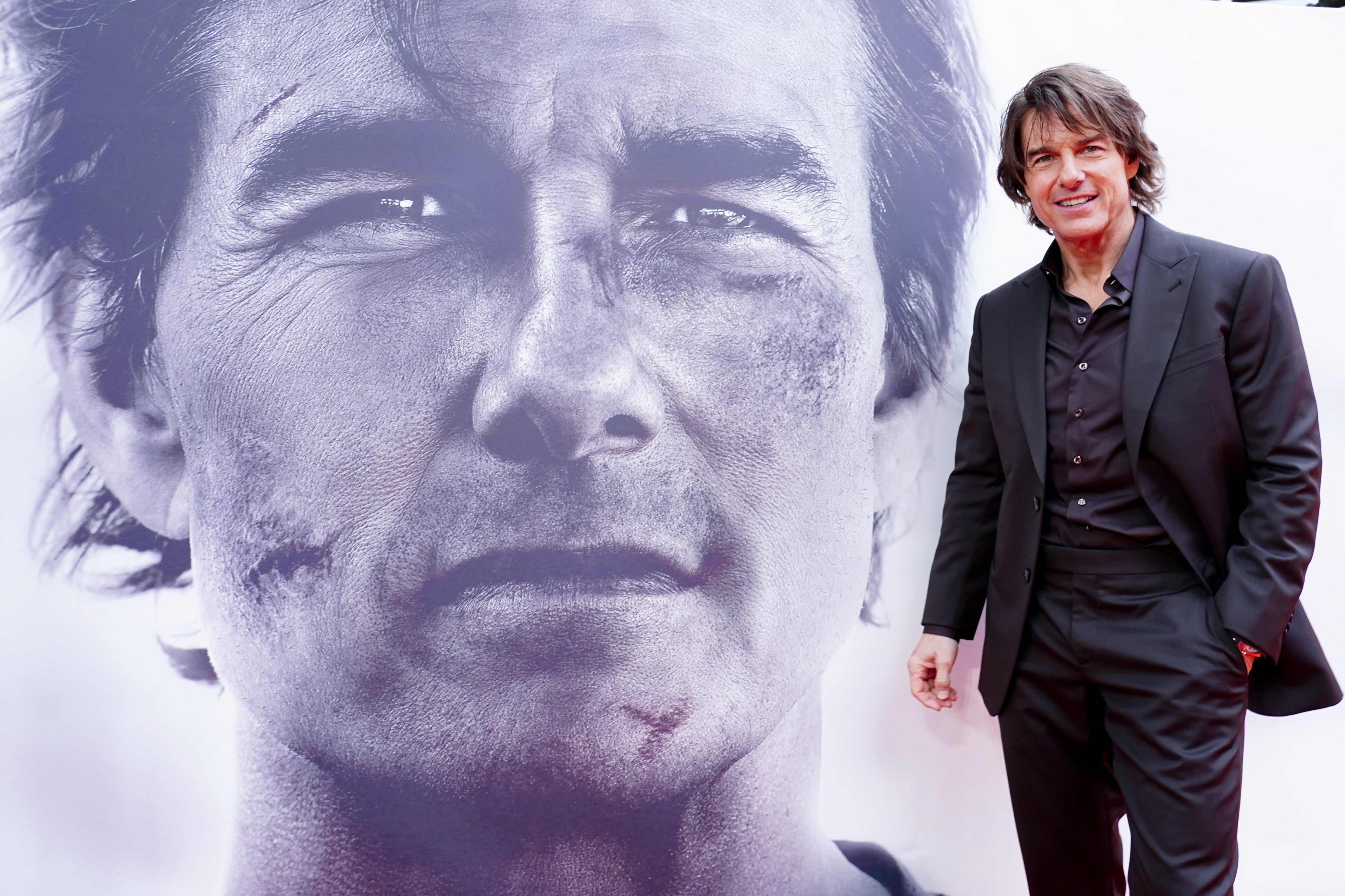A medium shot of Tom Cruise at a film premiere. He is dressed in a black suit and open-collared shirt, smiling at the camera. He stands to the right of a massive, black-and-white close-up banner of his own face, which shows him with weathered skin and a small scar on his cheek.