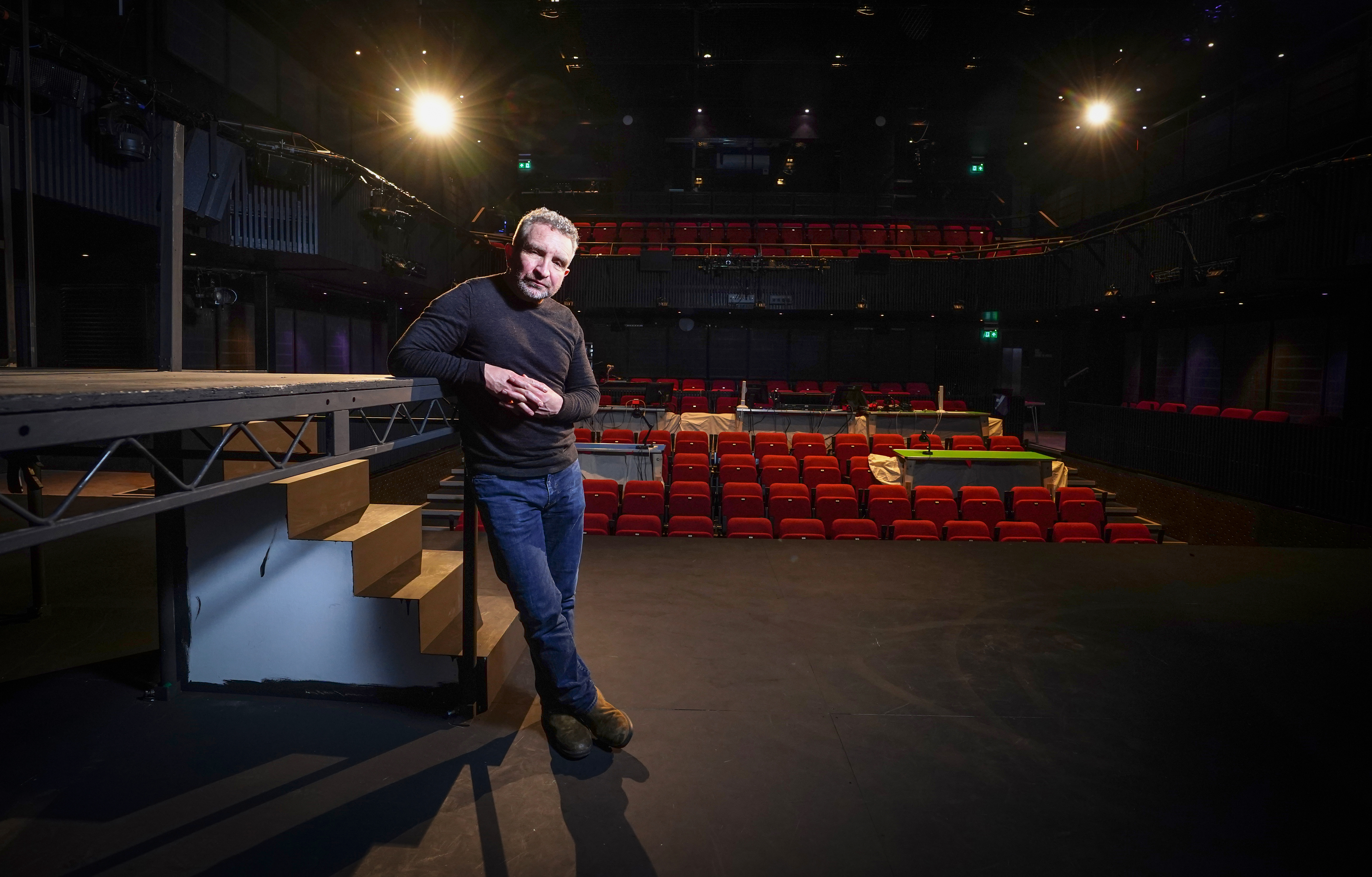 A wide-angle shot of actor Eddie Marsan on a darkened theater stage. He is dressed casually in a black long-sleeve shirt and jeans, leaning against a wooden set piece. Behind him, the empty red seats of the theater's stalls and balcony are visible under the glow of overhead stage lights.