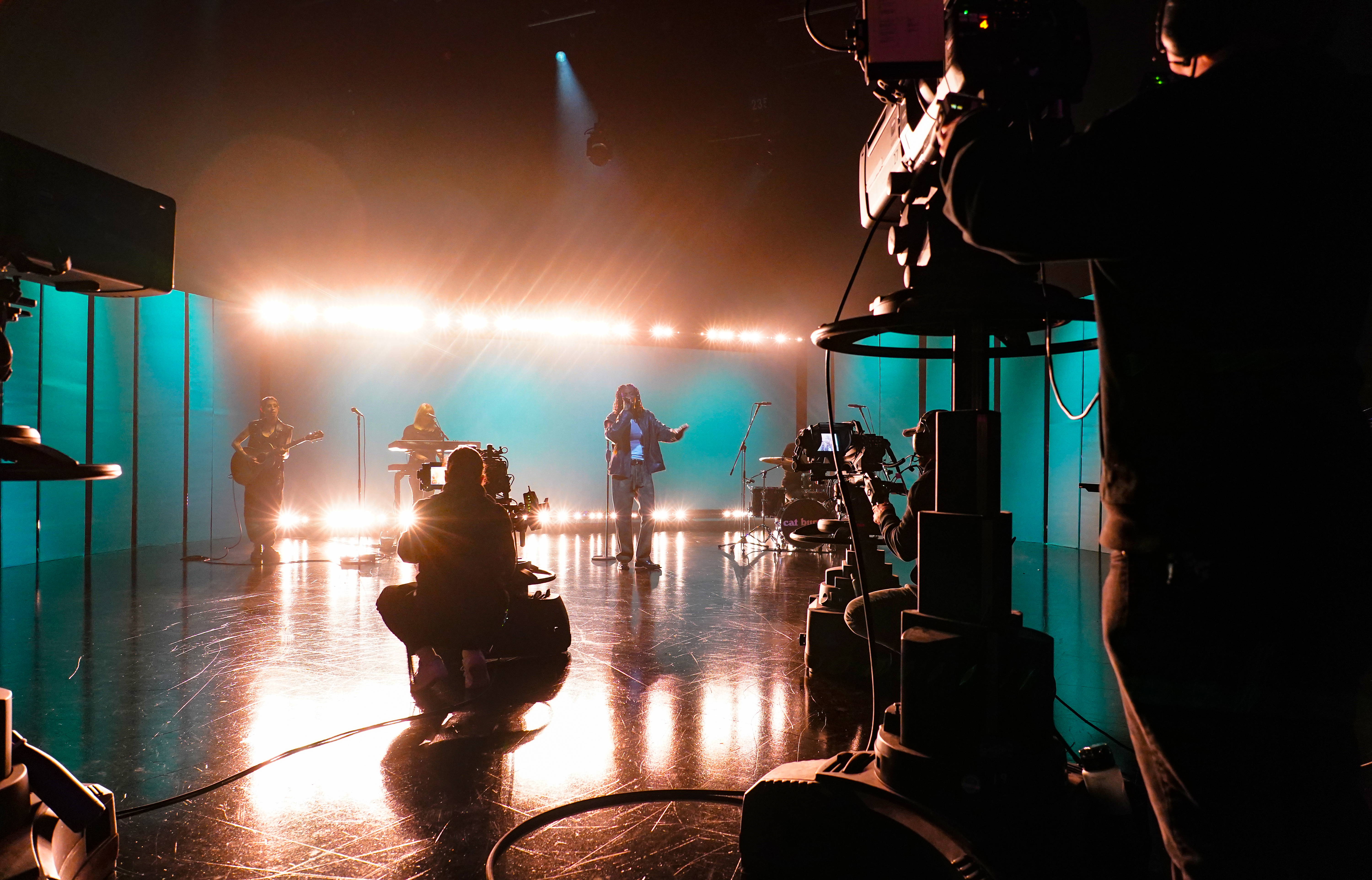 A wide shot of a television studio set where Cat Burns is performing with a live band. The stage is silhouetted against a bright, warm light bar and a teal-colored backdrop. In the foreground, the silhouettes of professional television cameras and operators are visible, framing the performance.