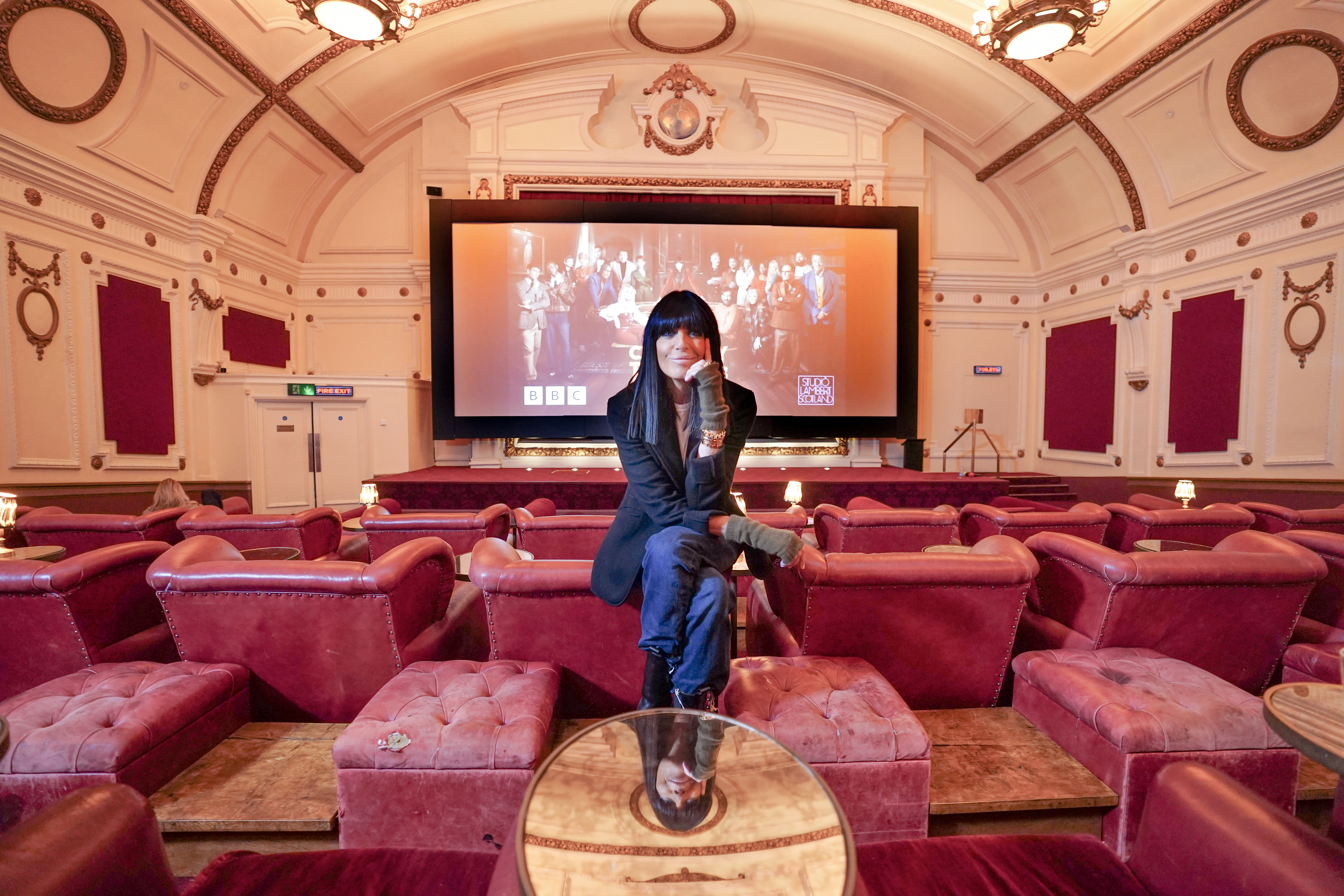 Claudia Winkleman rests her chin on her hand as she sits in a classic cinema. The traitors is on the screen