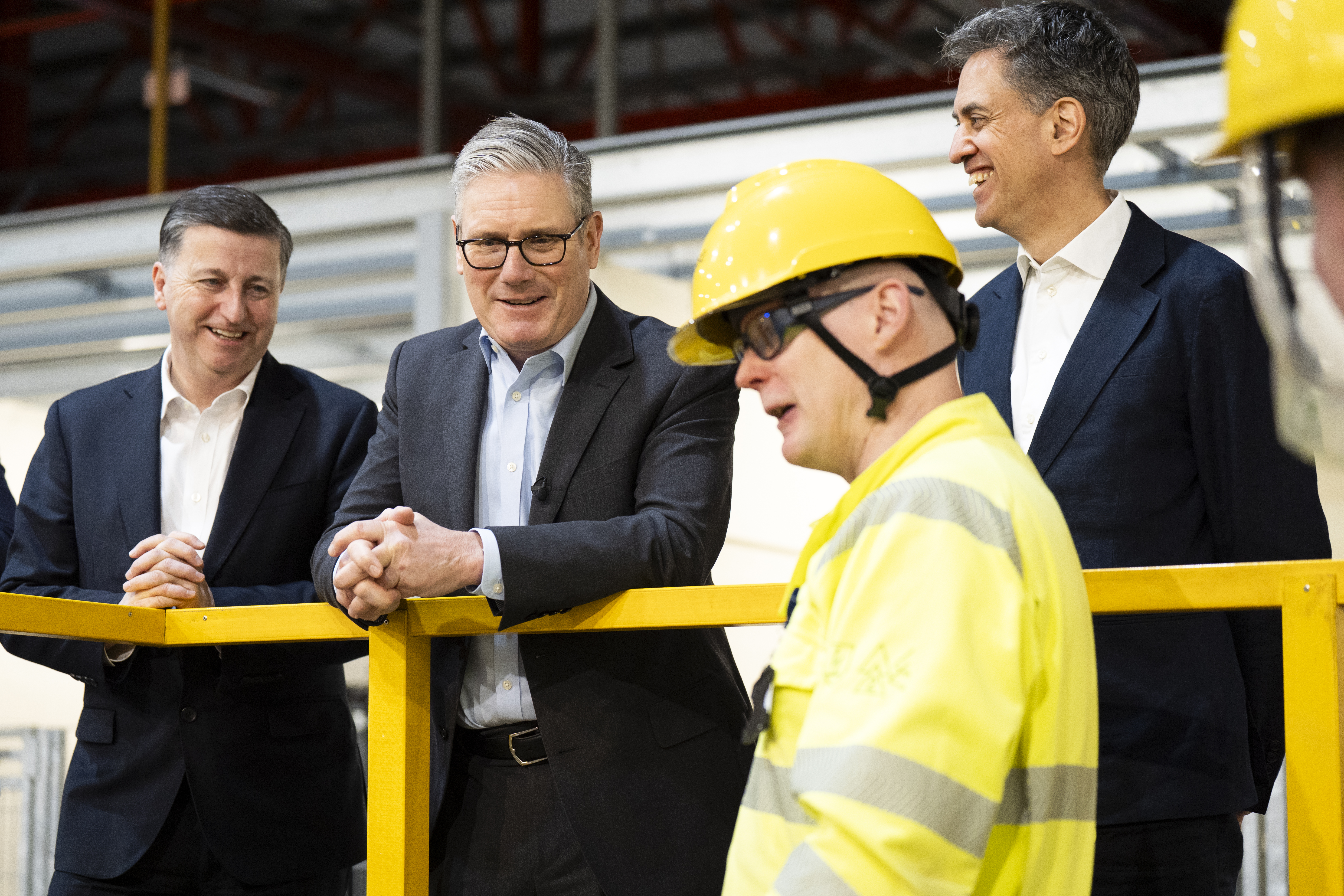Douglas Alexander, Sir Keir Starmer and Ed Miliband smiling while being addressed by a man in hi-vis clothing