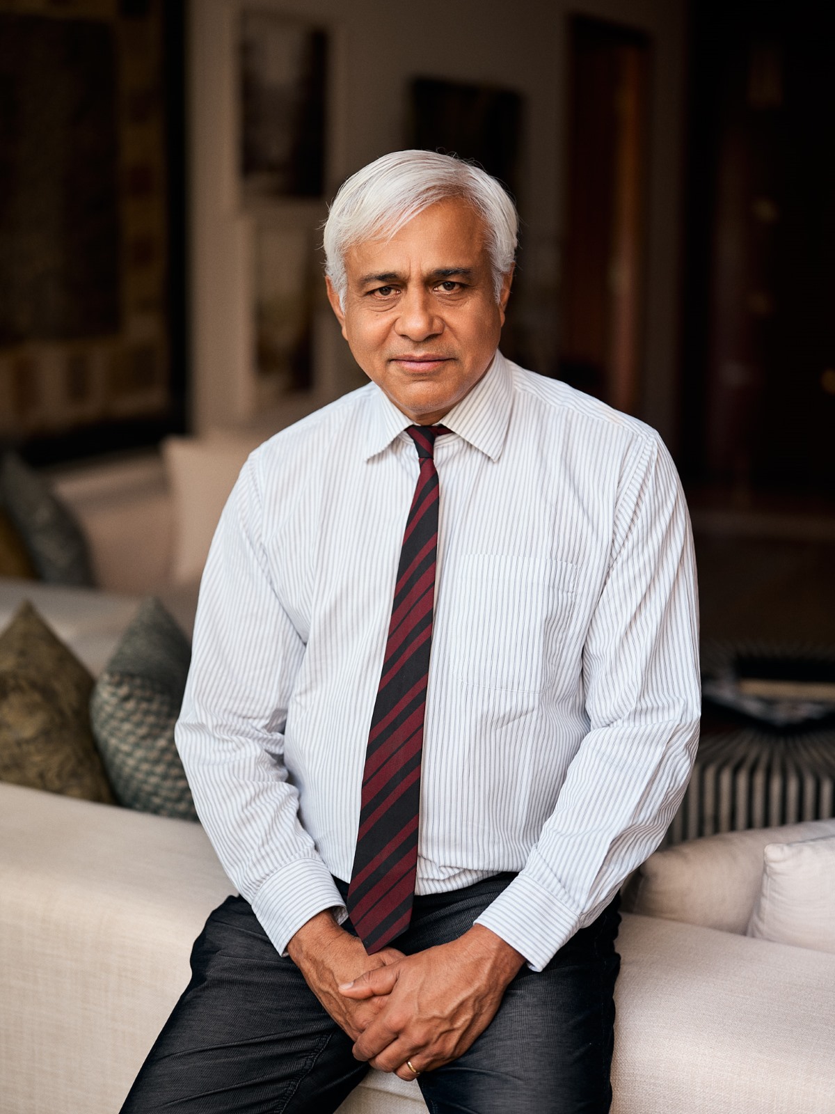 Handout photo of Mahesh Aditya, seated on the back of a sofa wearing a shirt and tie