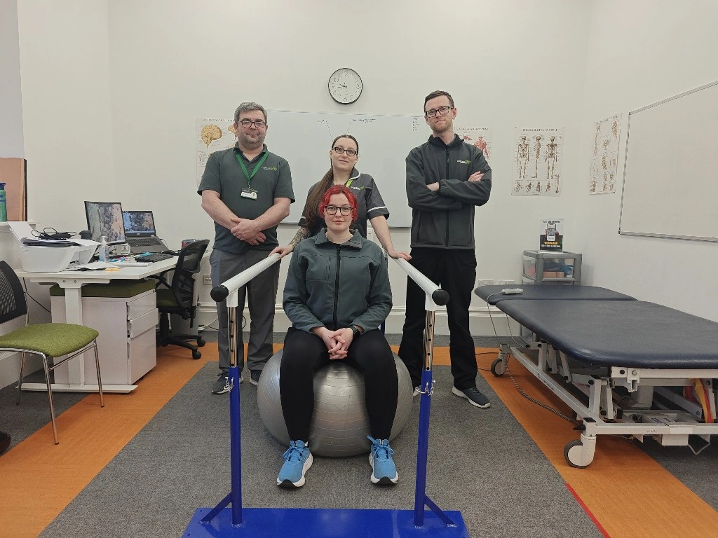 Two men and a woman sitting behind a woman sitting on a physiotherapy ball in a clinic setting