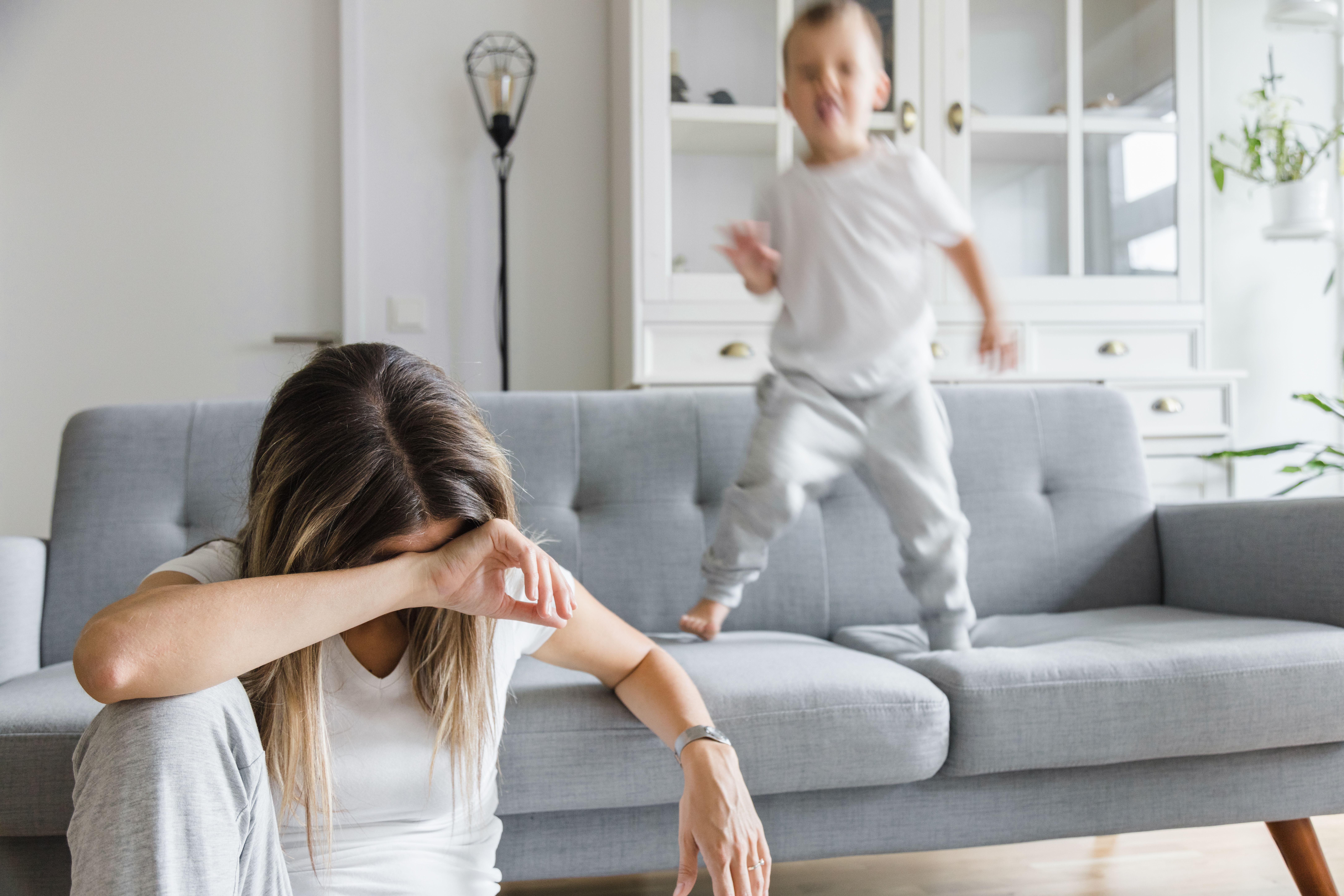A tired mother rests her head on her hand while sitting on the floor while a child bounces on a sofa behind her