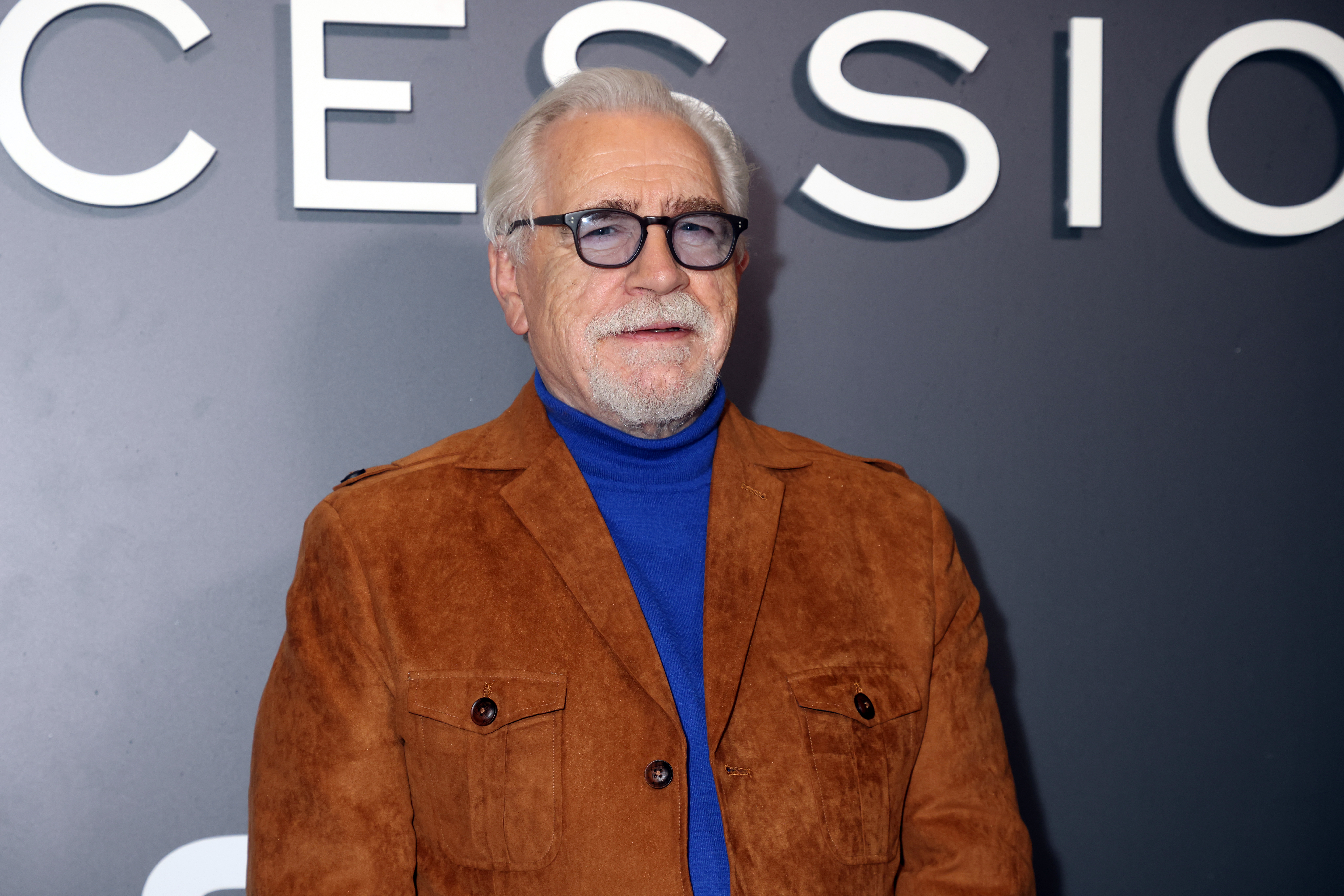 Brian Cox posing for photos in a brown jacket and blue top at a red carpet event