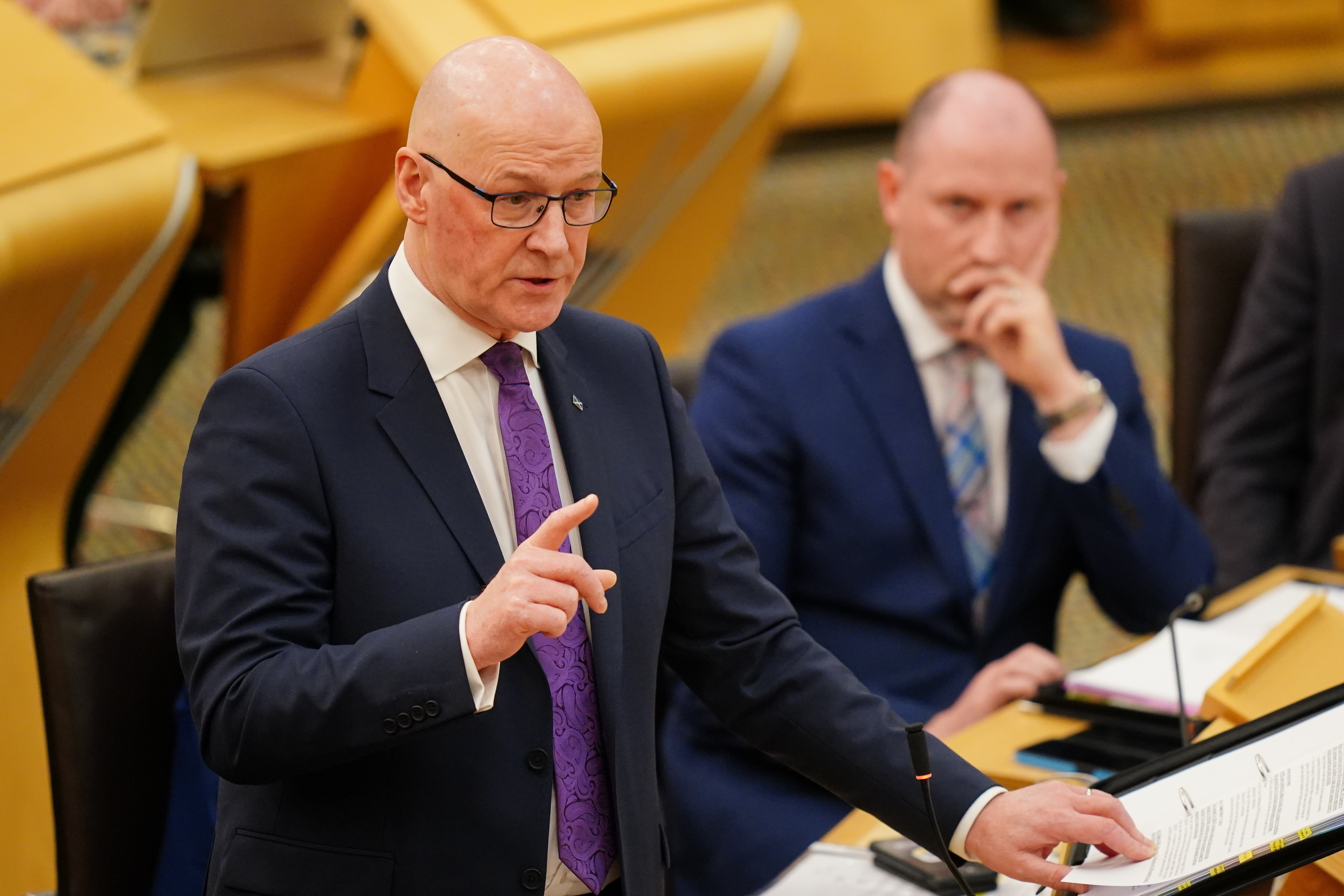 John Swinney standing while speaking in Holyrood, with Neil Gray seated beside him