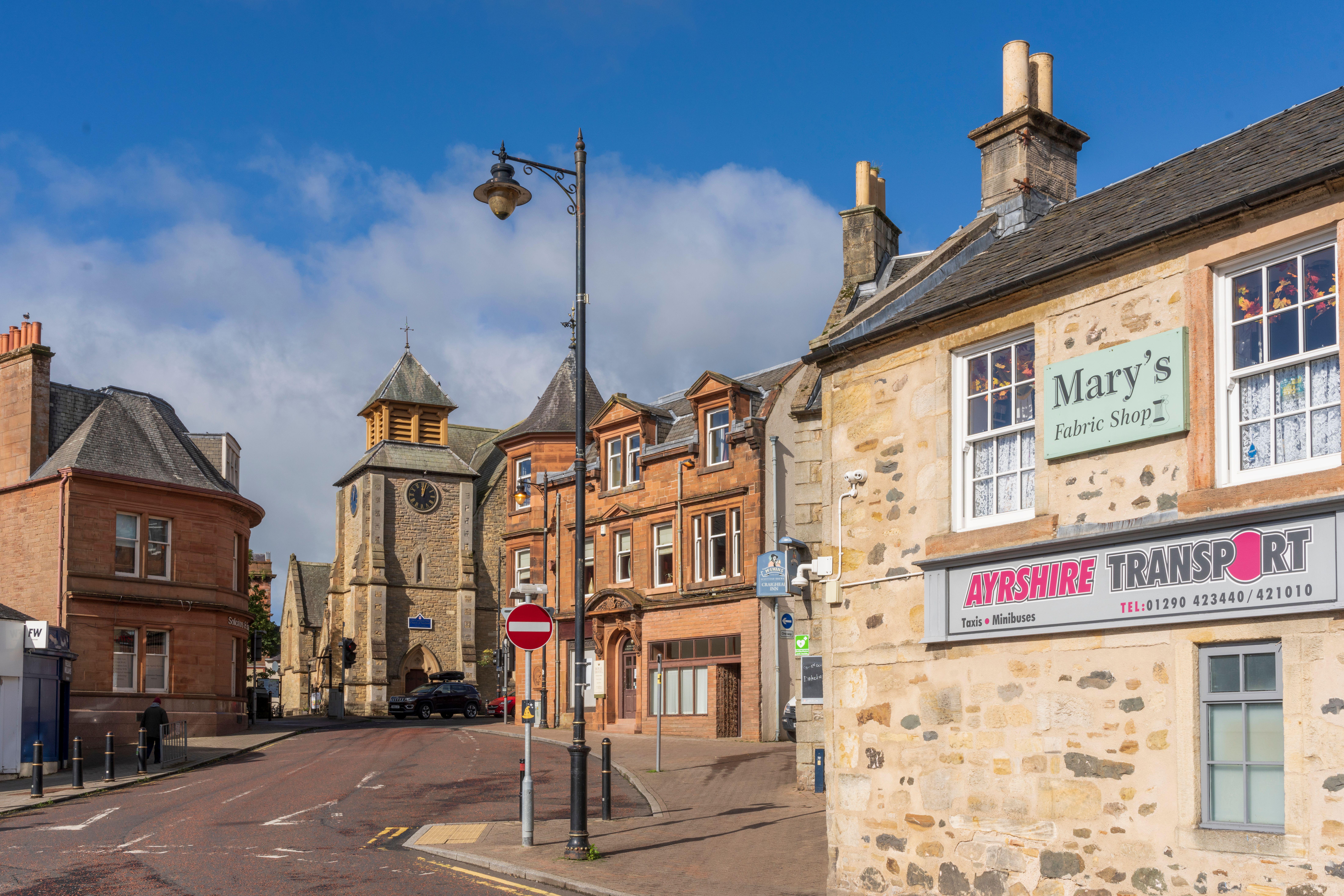 A sunny Scottish town centre featuring a clock tower, stone buildings, and street life with shops and signage.