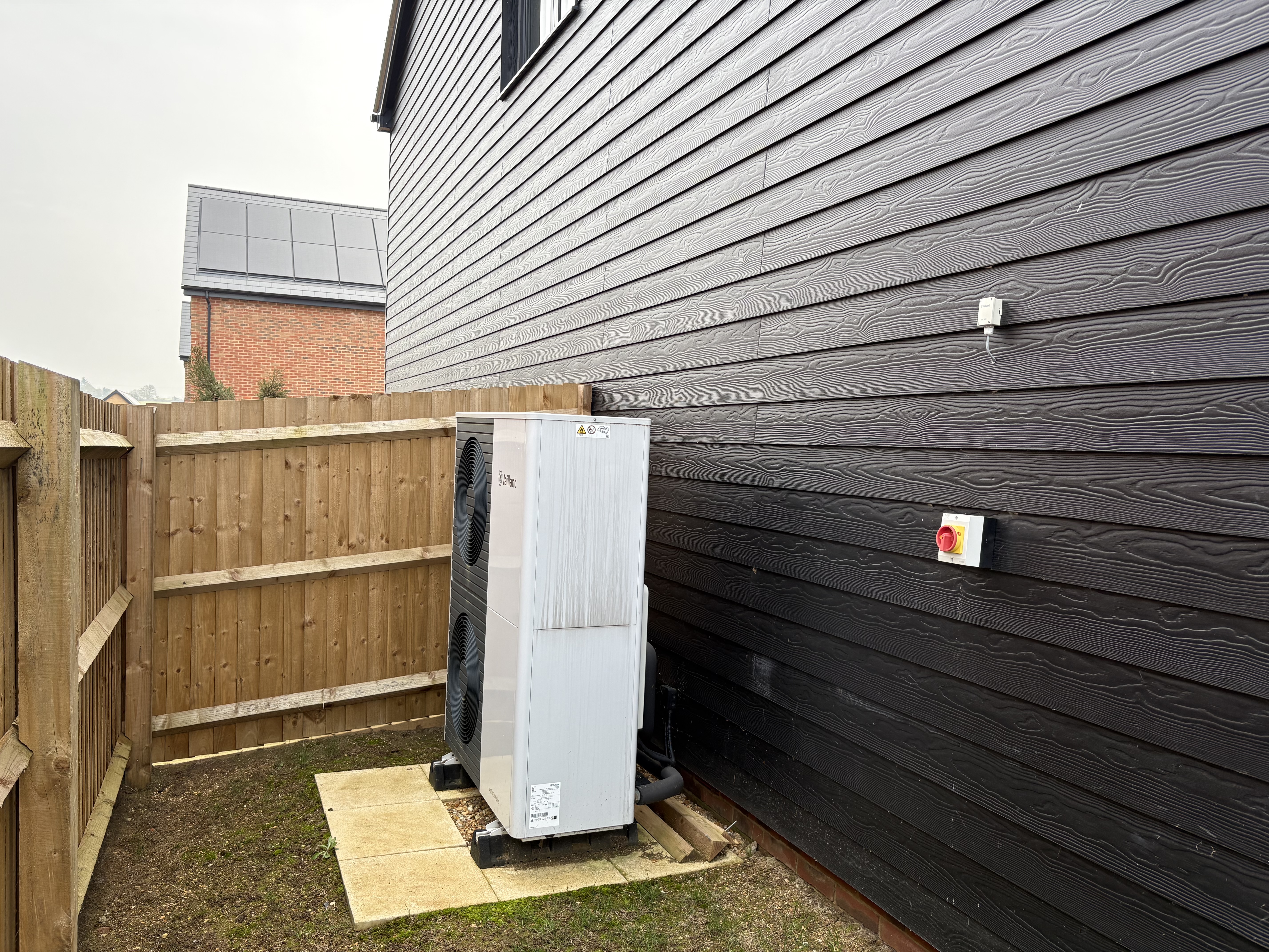 A heat pump against the side of a black clapboard style cladded wall of a home surrounded by fencing