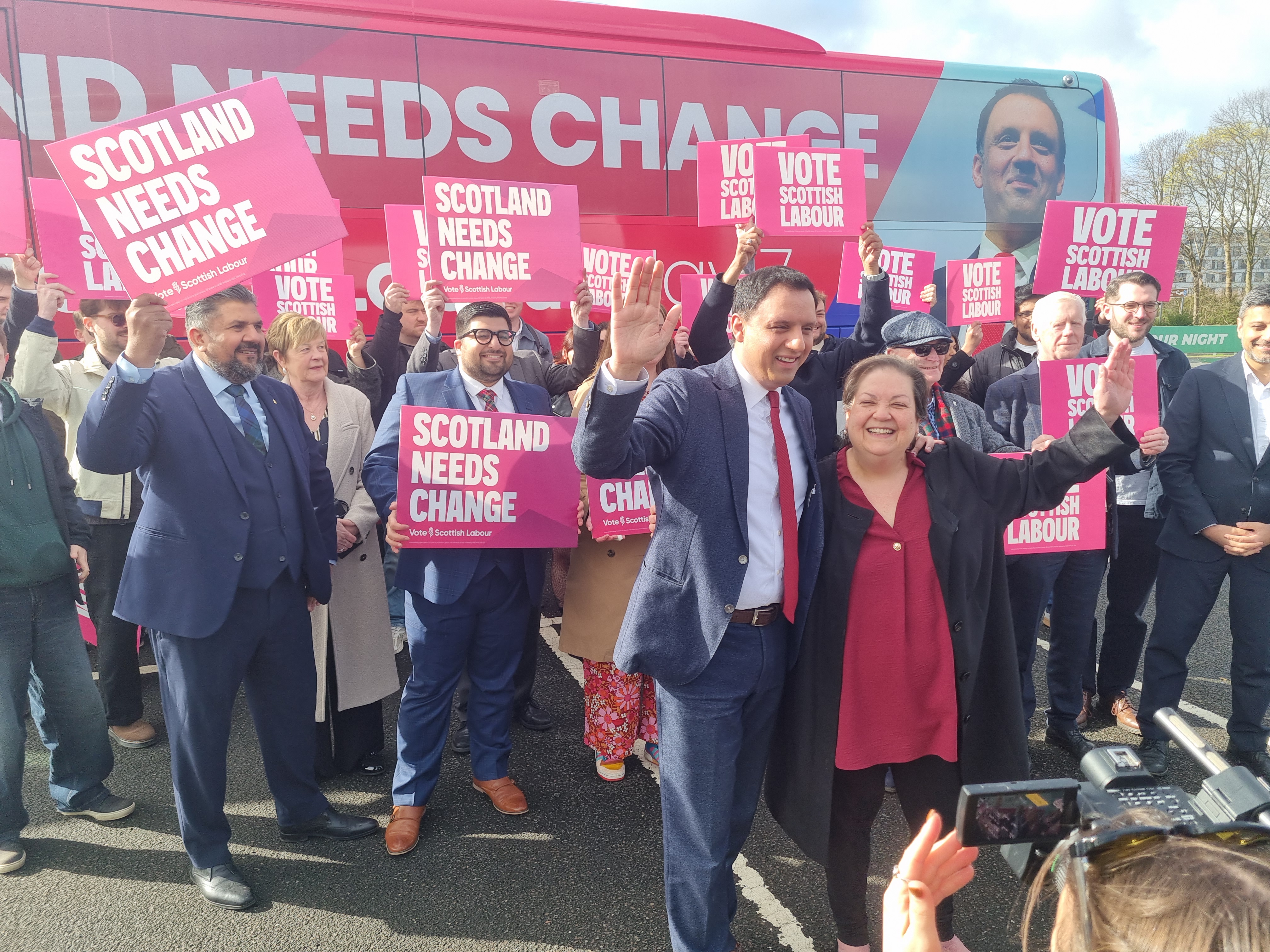 Anas Sarwar and Dame Jackie Baillie smiling and waving amongst Labour supporters in front of the party's battle bus