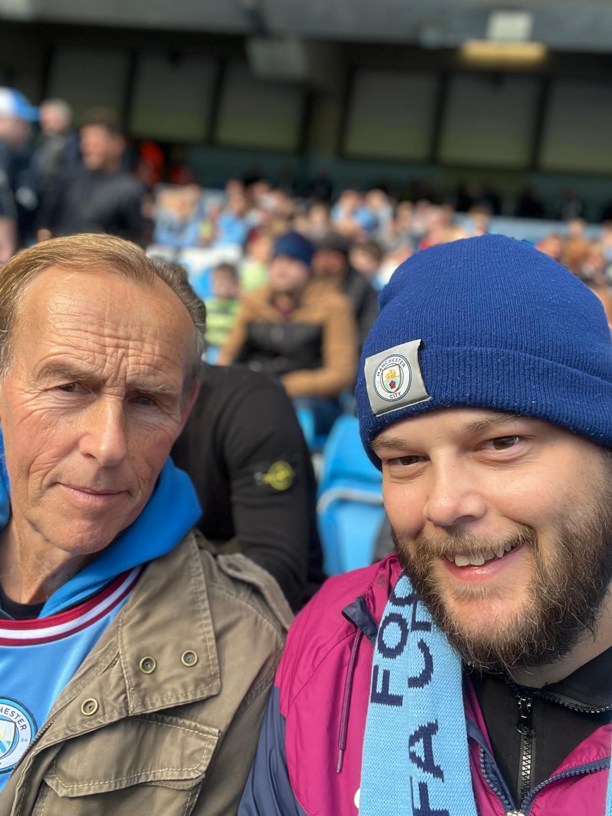 Neil Howarth, wearing a Manchester City hat and scarf, in a football stadium stand with his father Peter, who is wearing a Manchester City shirt