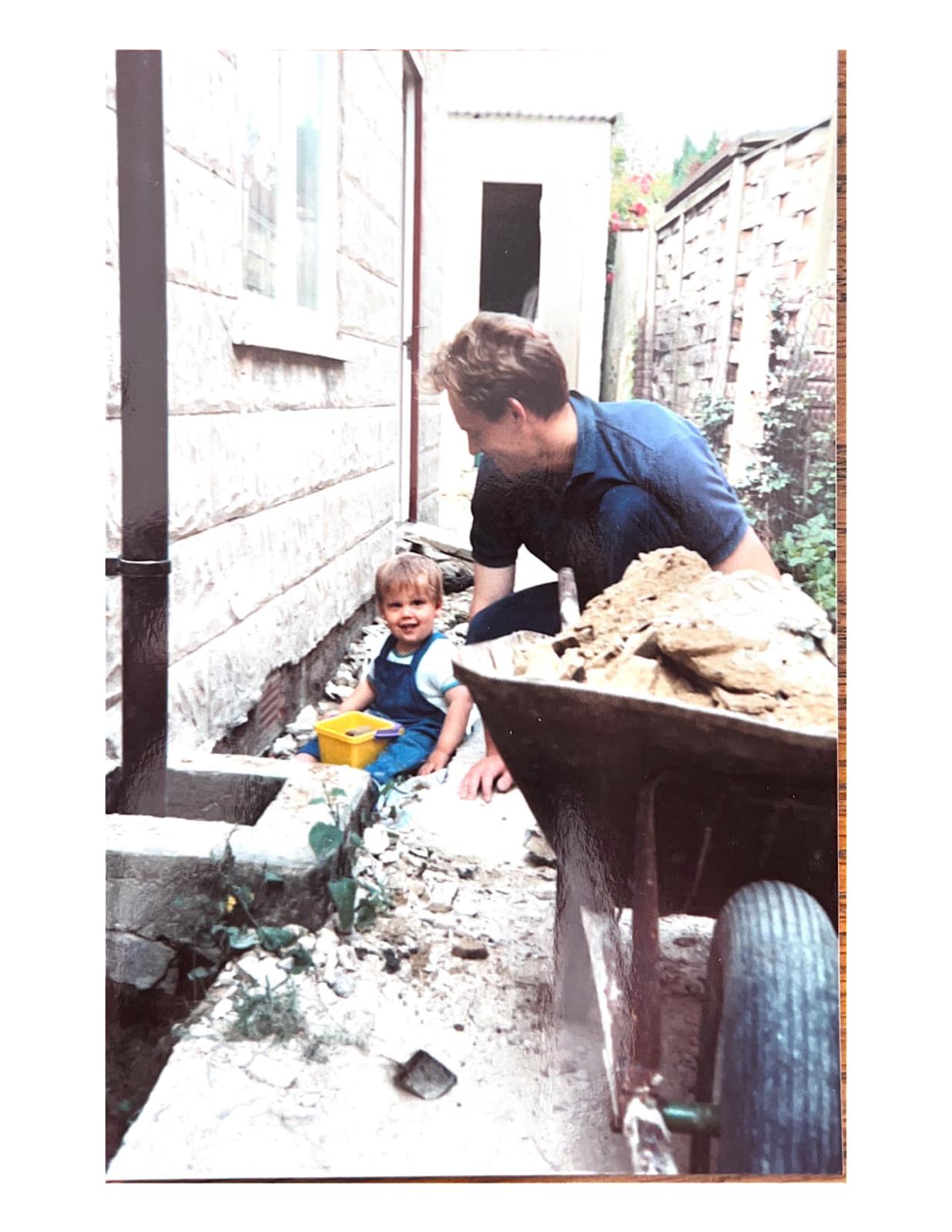 Neil as a young boy sitting on the ground with a yellow bucket next to his dad, Peter, who has a wheelbarrow full of rubble