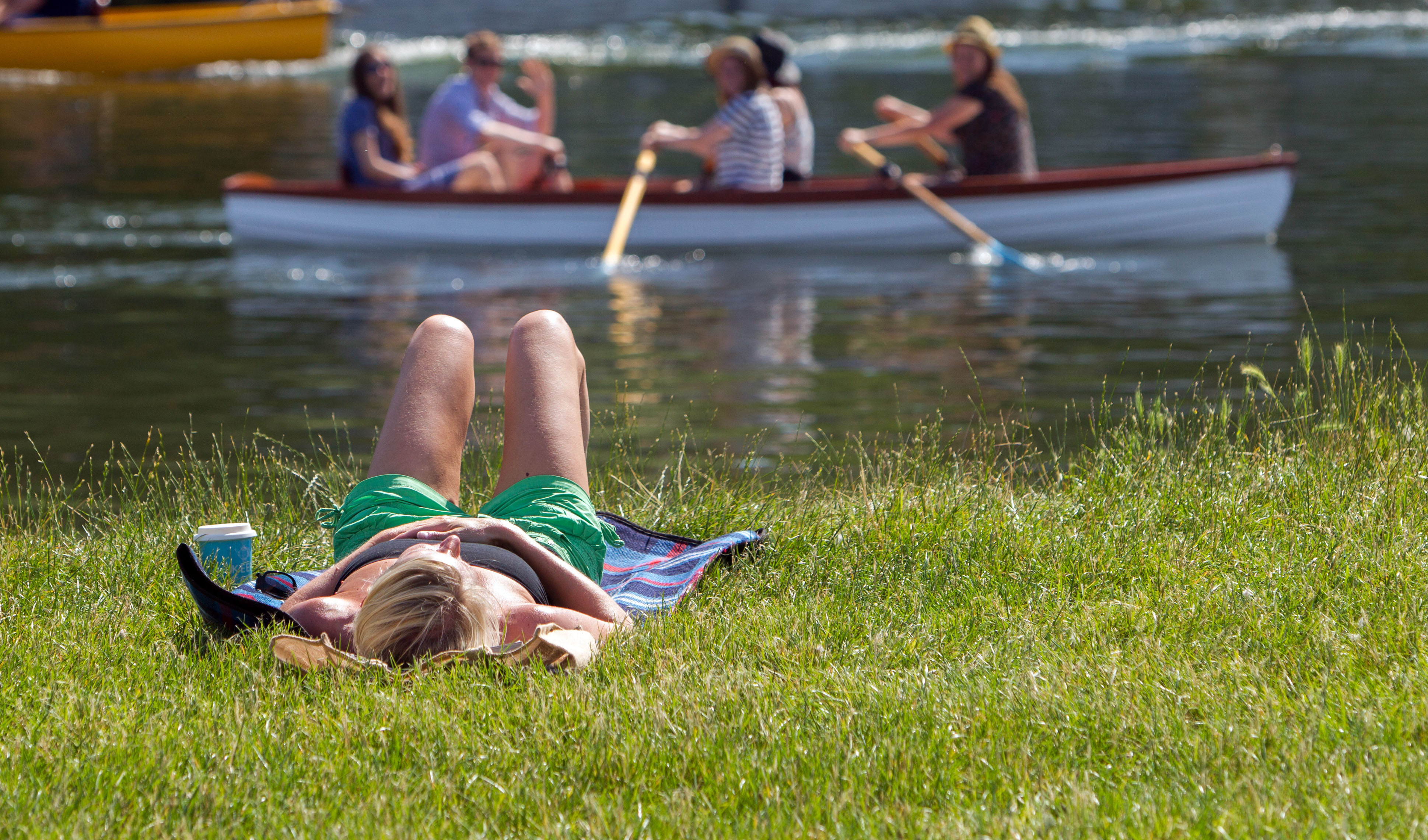 A woman sunbathing on grass, with people in a row boat in the background