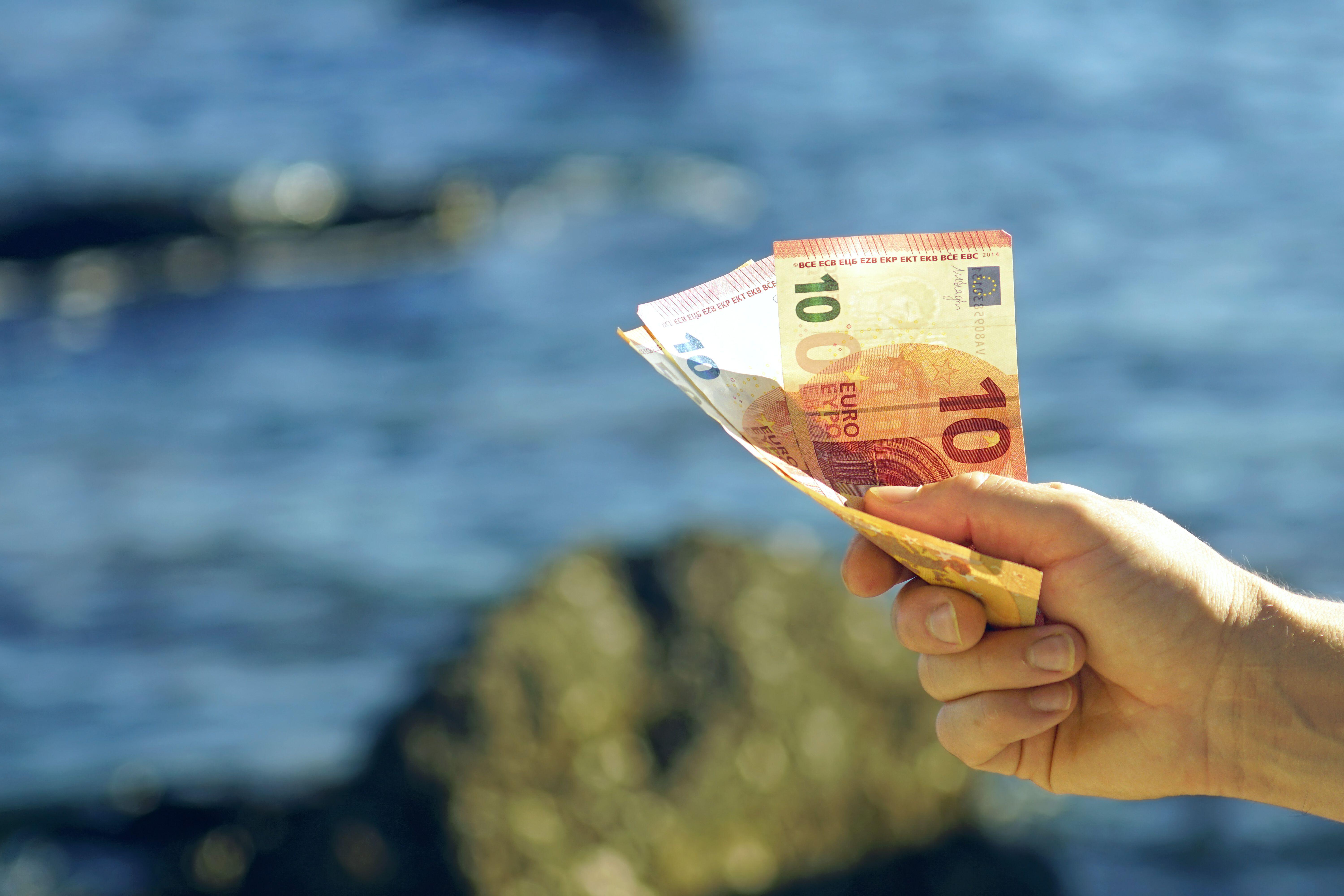 A hand clutching some euro notes in the foreground, with the sea shown in the distance