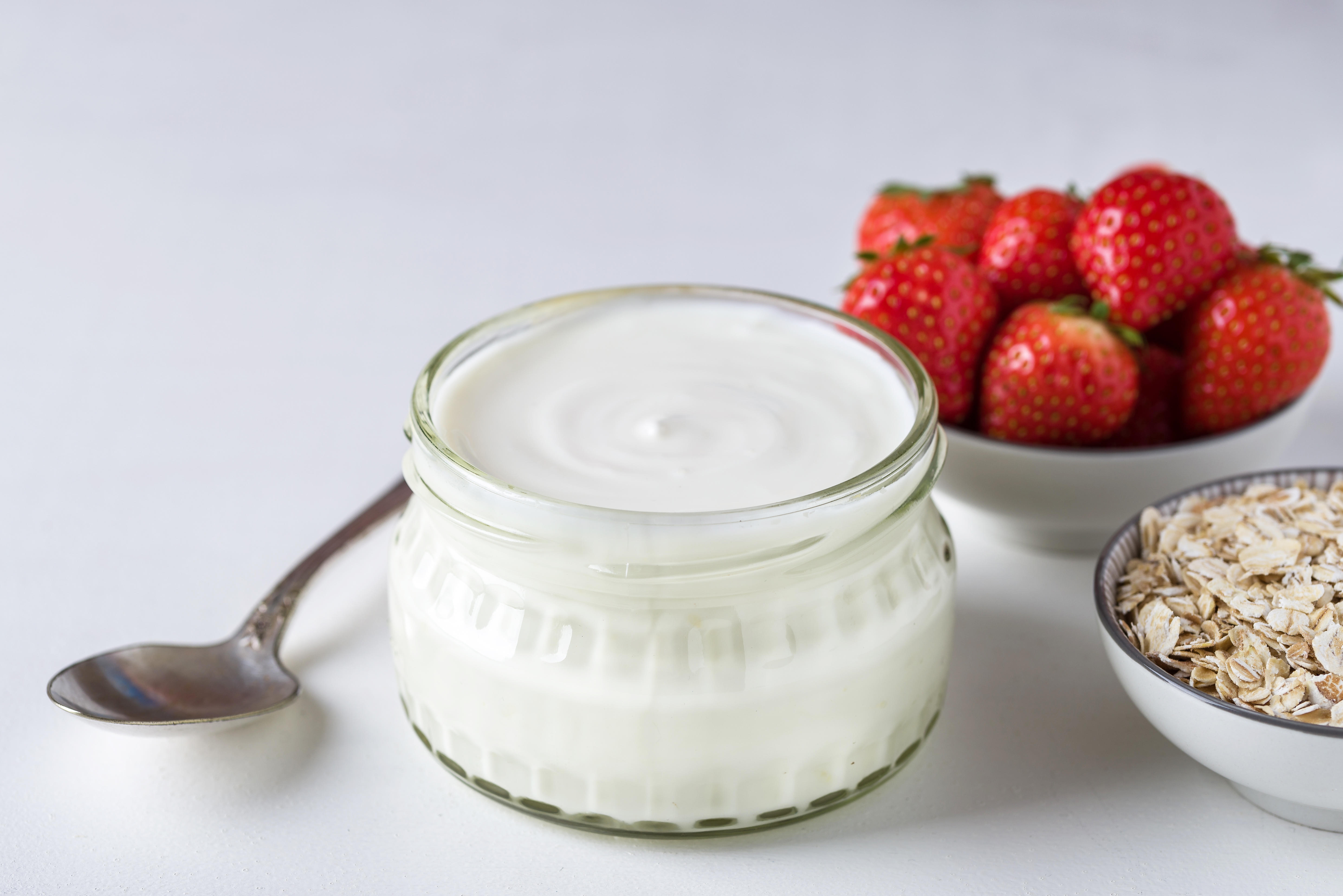 White yogurt in glass bowl next to a spoon, some oats and strawberries 