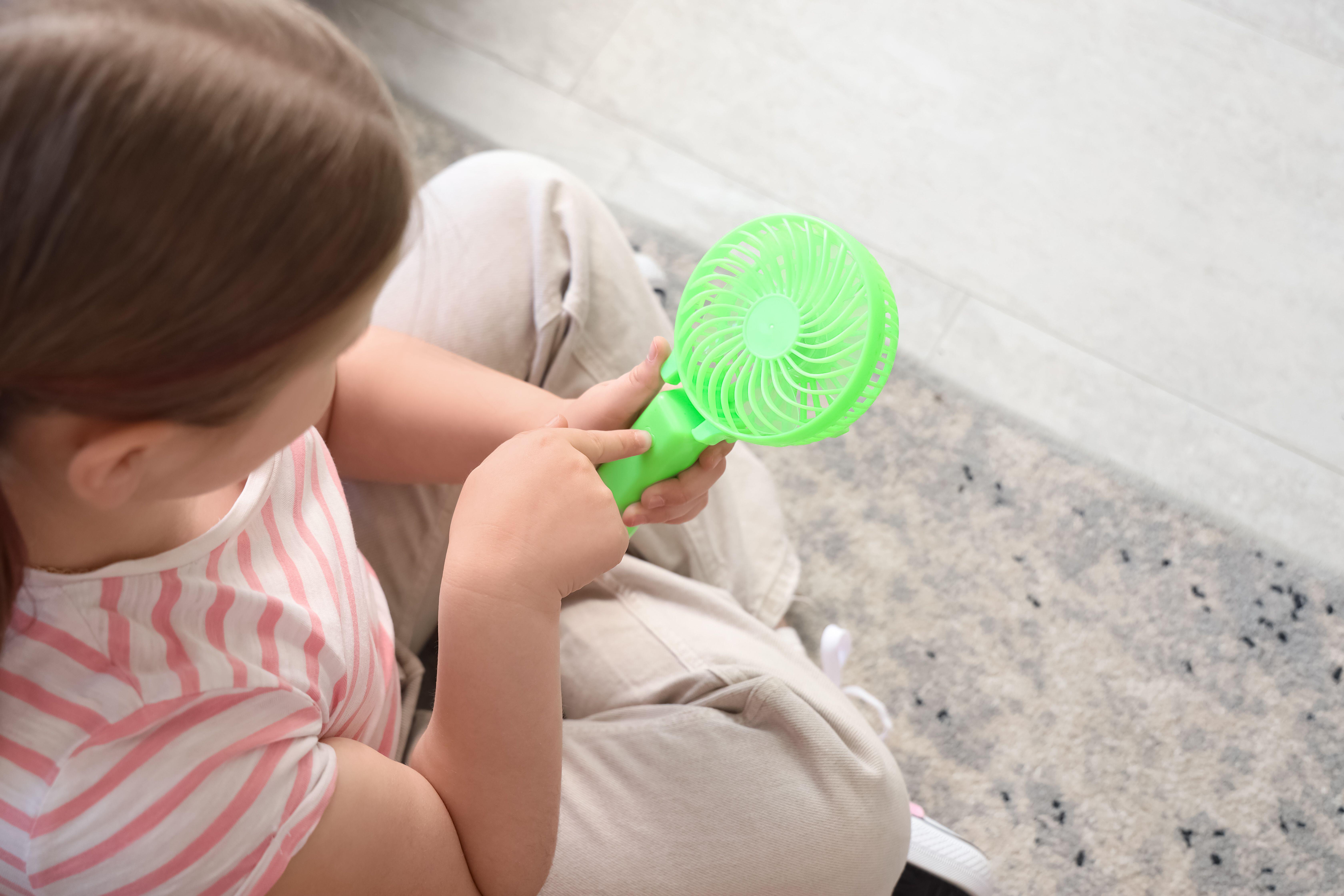 A young girl turning on a mini fan while sitting on the carpet at home