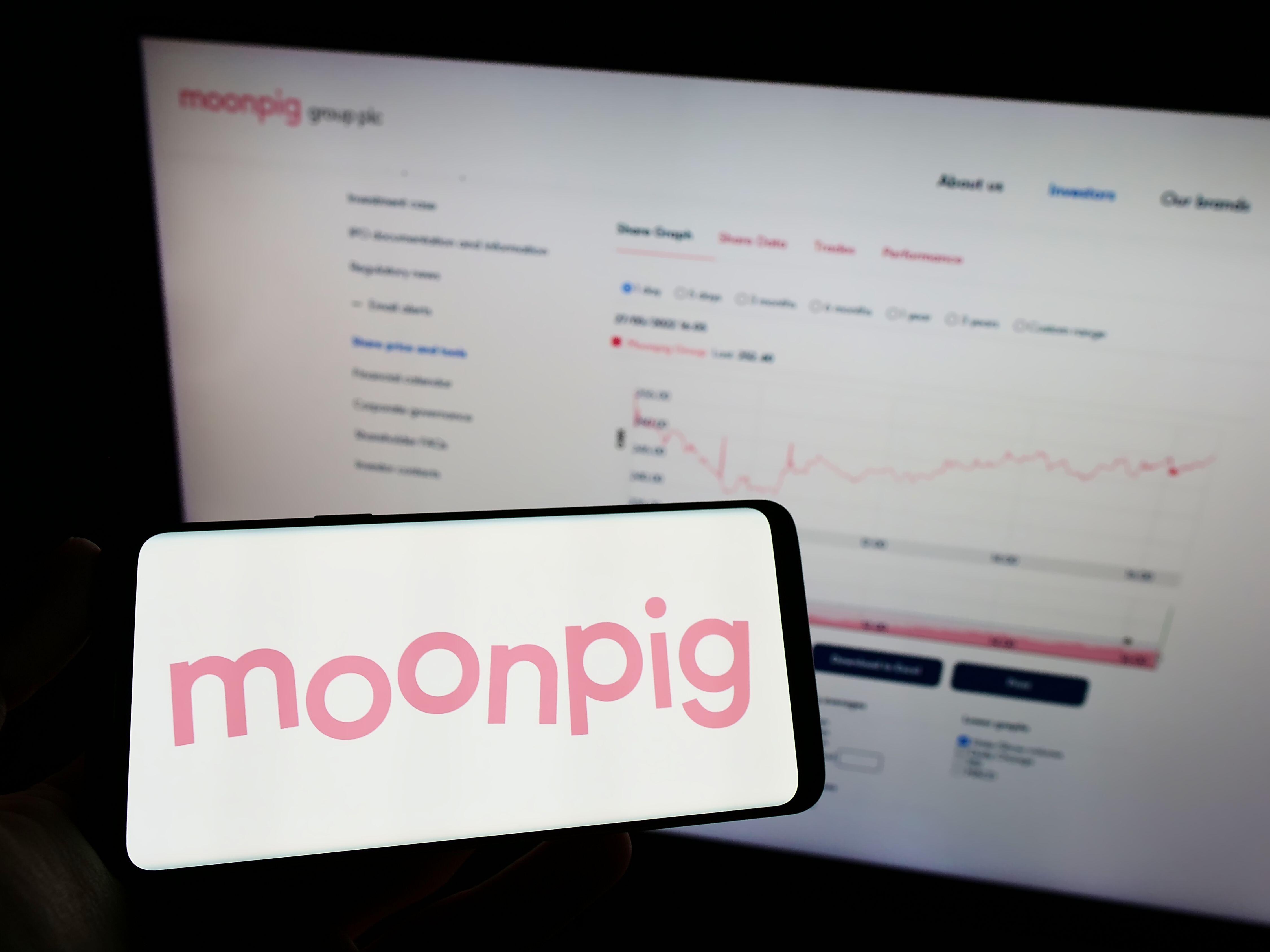 Person holding mobile phone with logo of Moonpig on screen in front of business web page