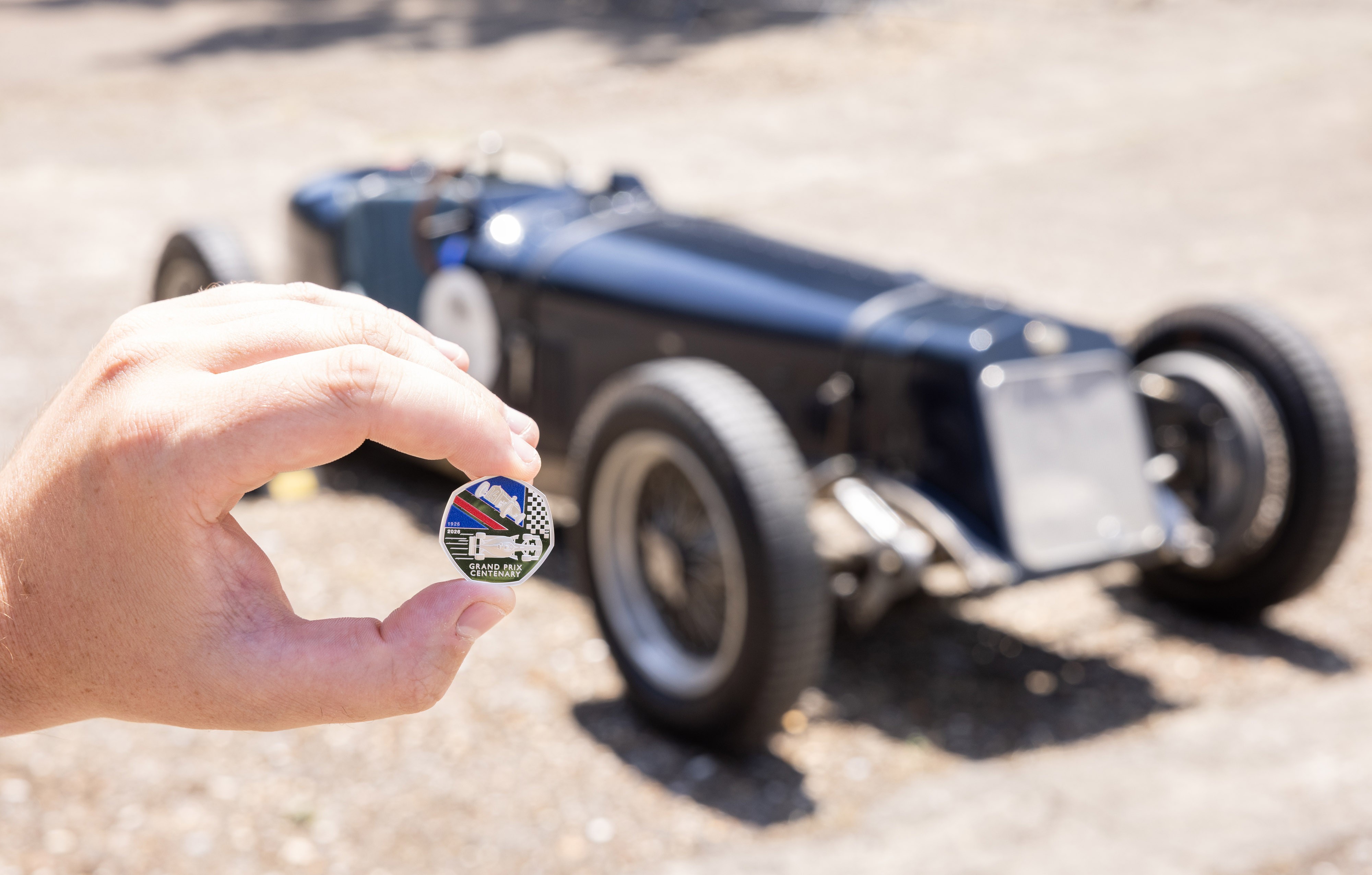 A hand holding a new 50p coin, in front of a navy Delage car